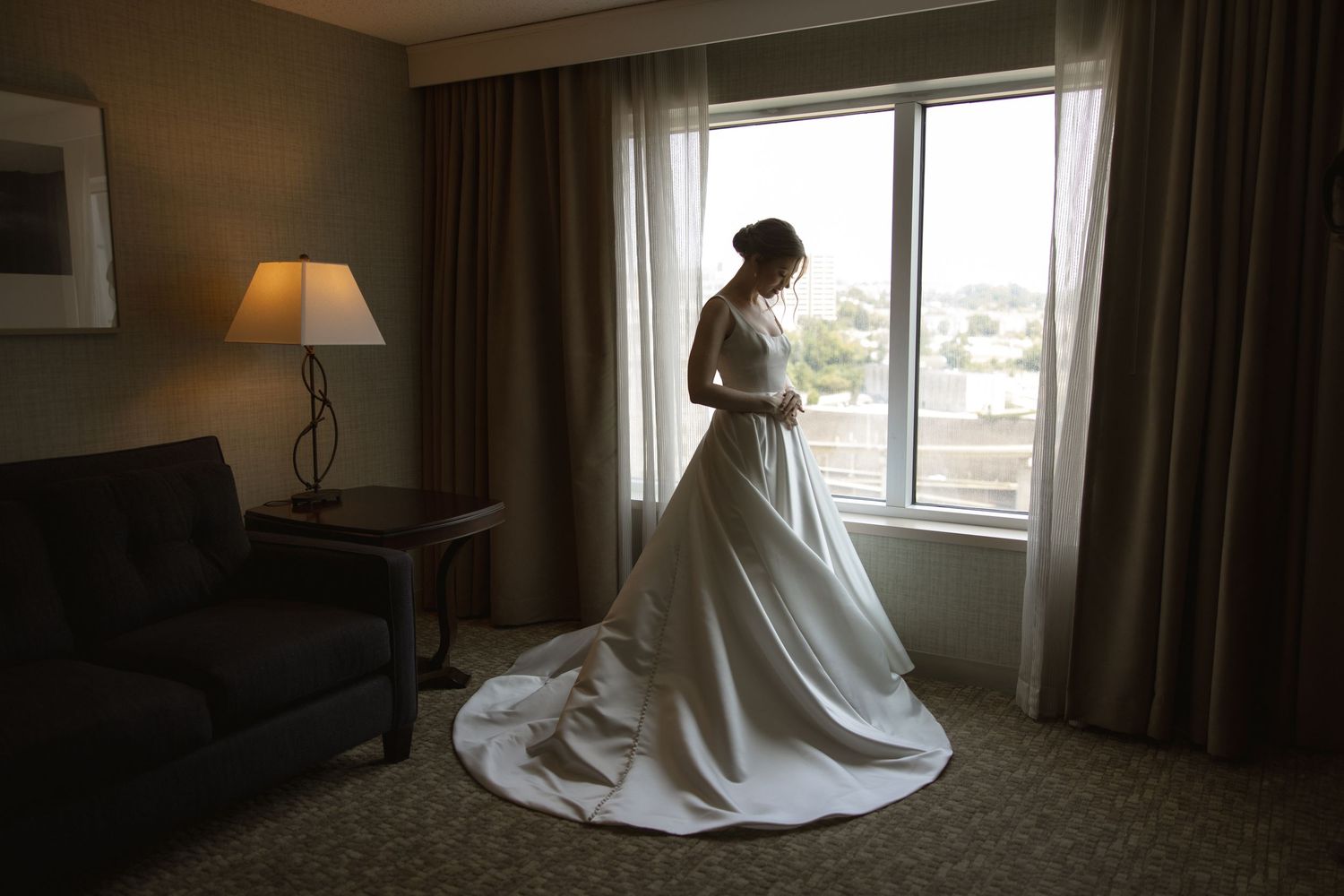 Silhouette of bride in flowing wedding gown standing by hotel room window.