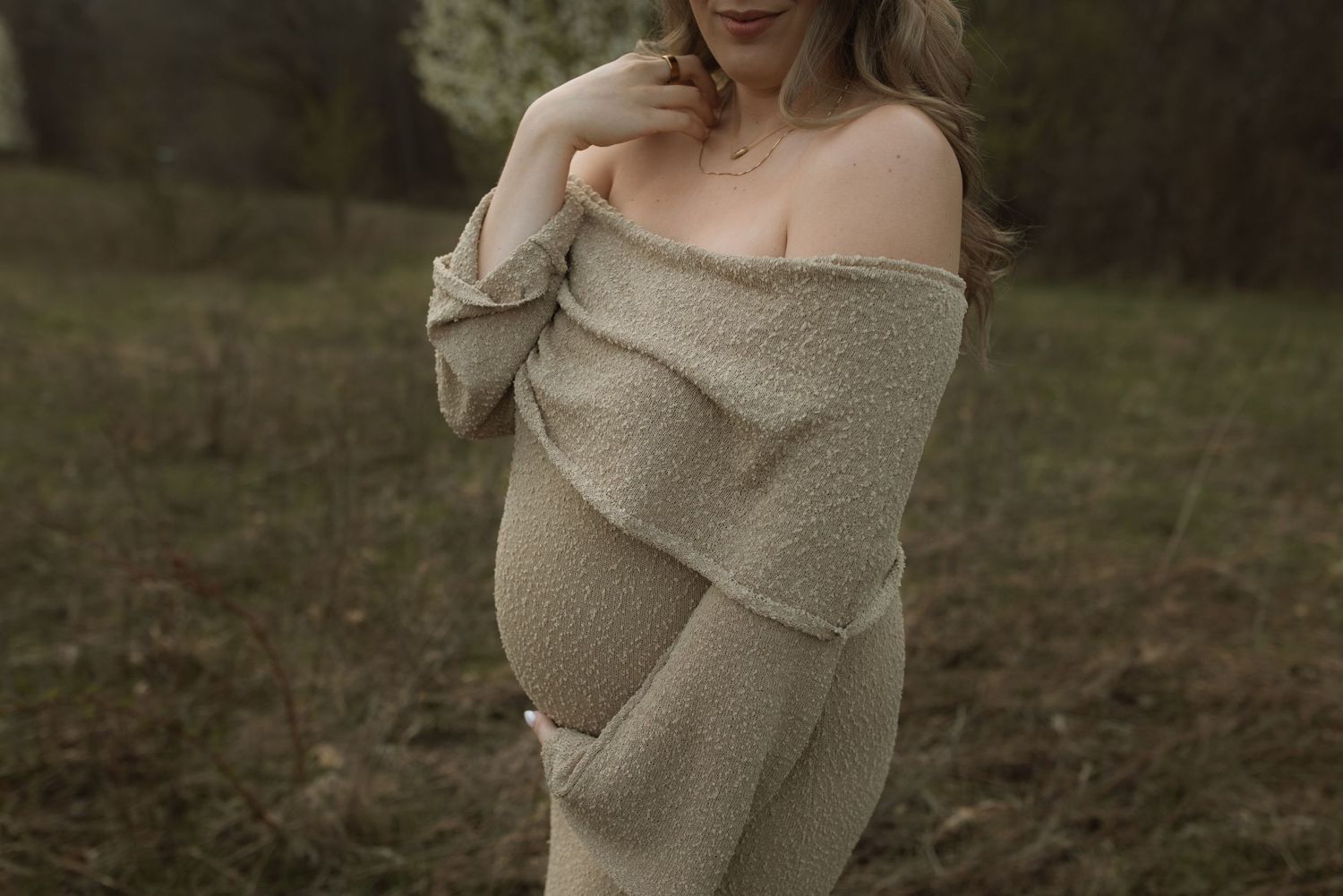 Maternity portrait featuring off-shoulder beige knit dress in natural setting.