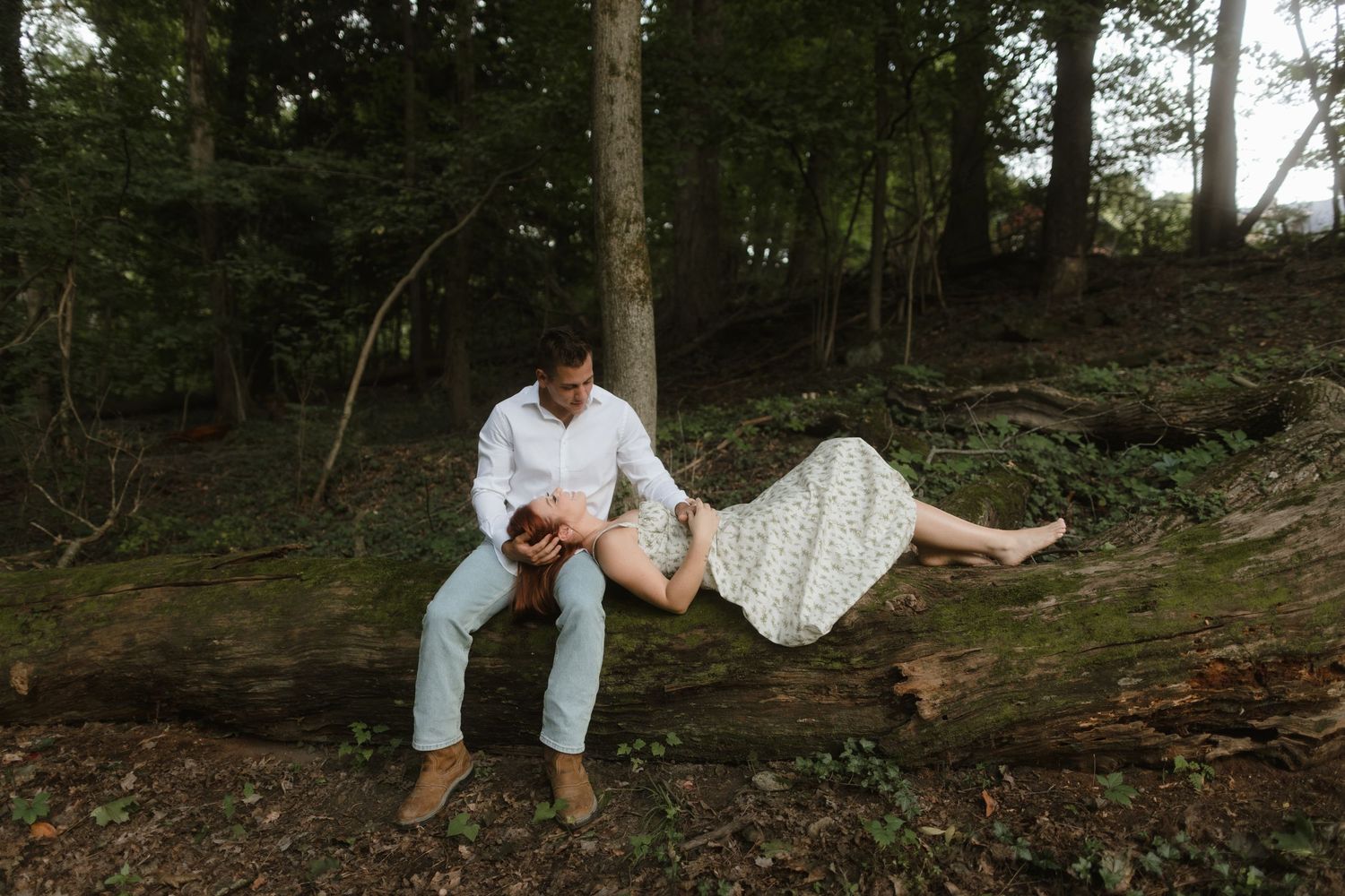 A couple in casual attire sits together on a fallen tree trunk in a wooded forest setting.