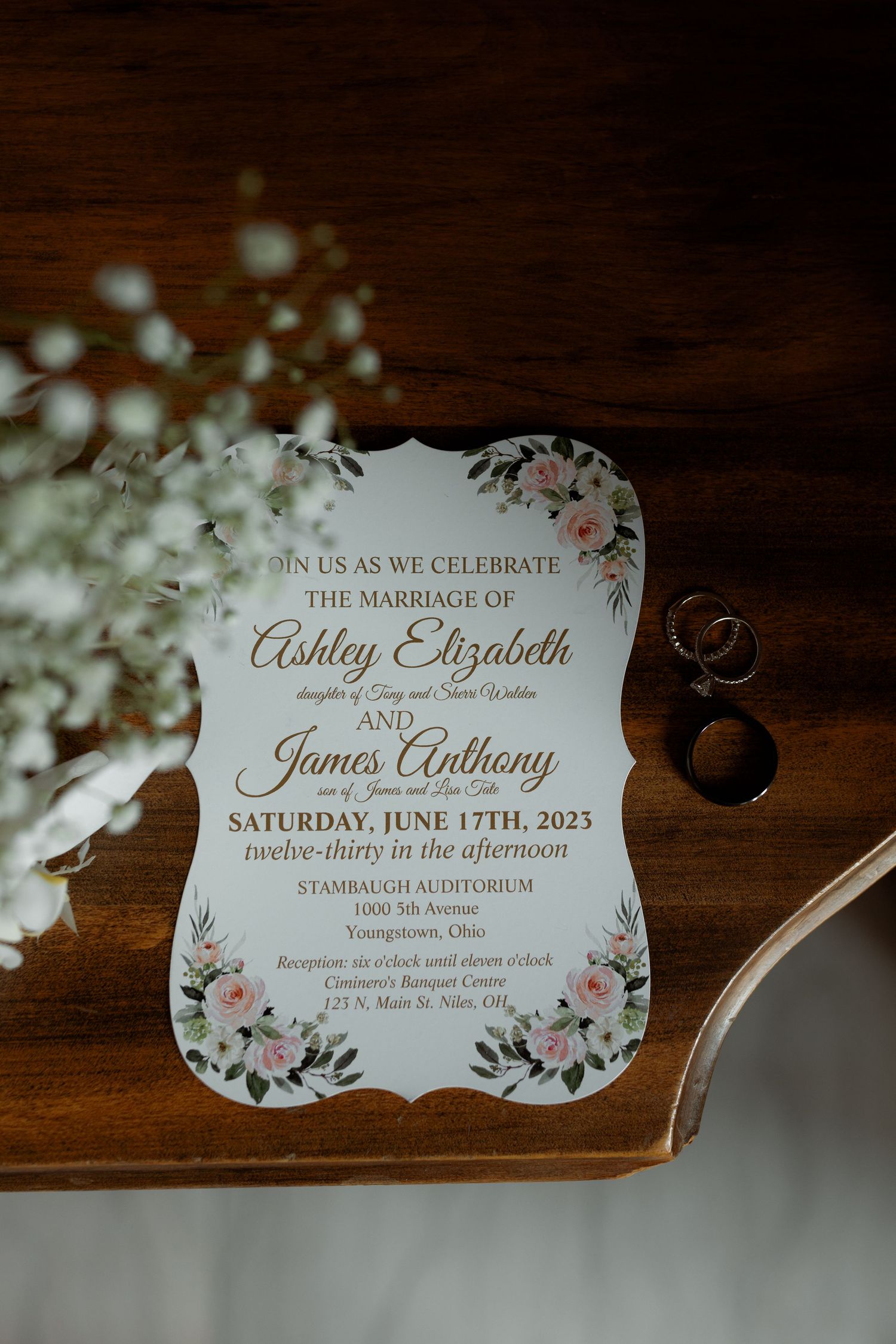 Wedding invitation with floral design and elegant calligraphy text on dark wood surface with rings beside it.