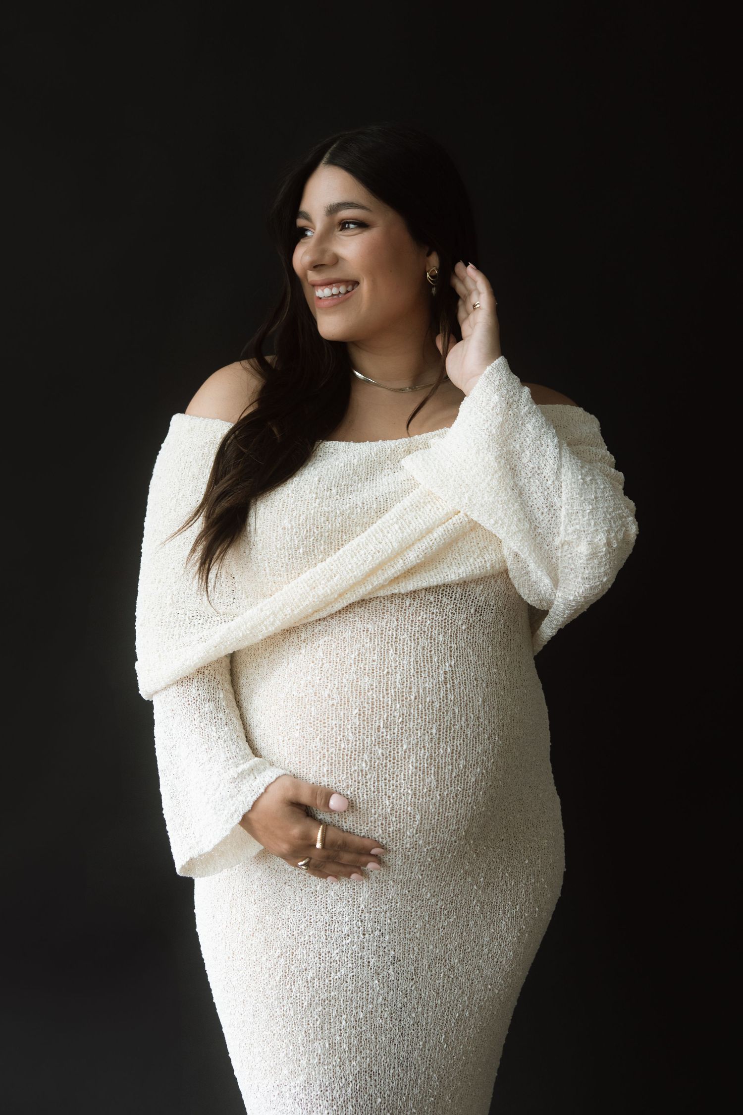 A maternity photo session featuring a white knit wrap dress against a dark background.