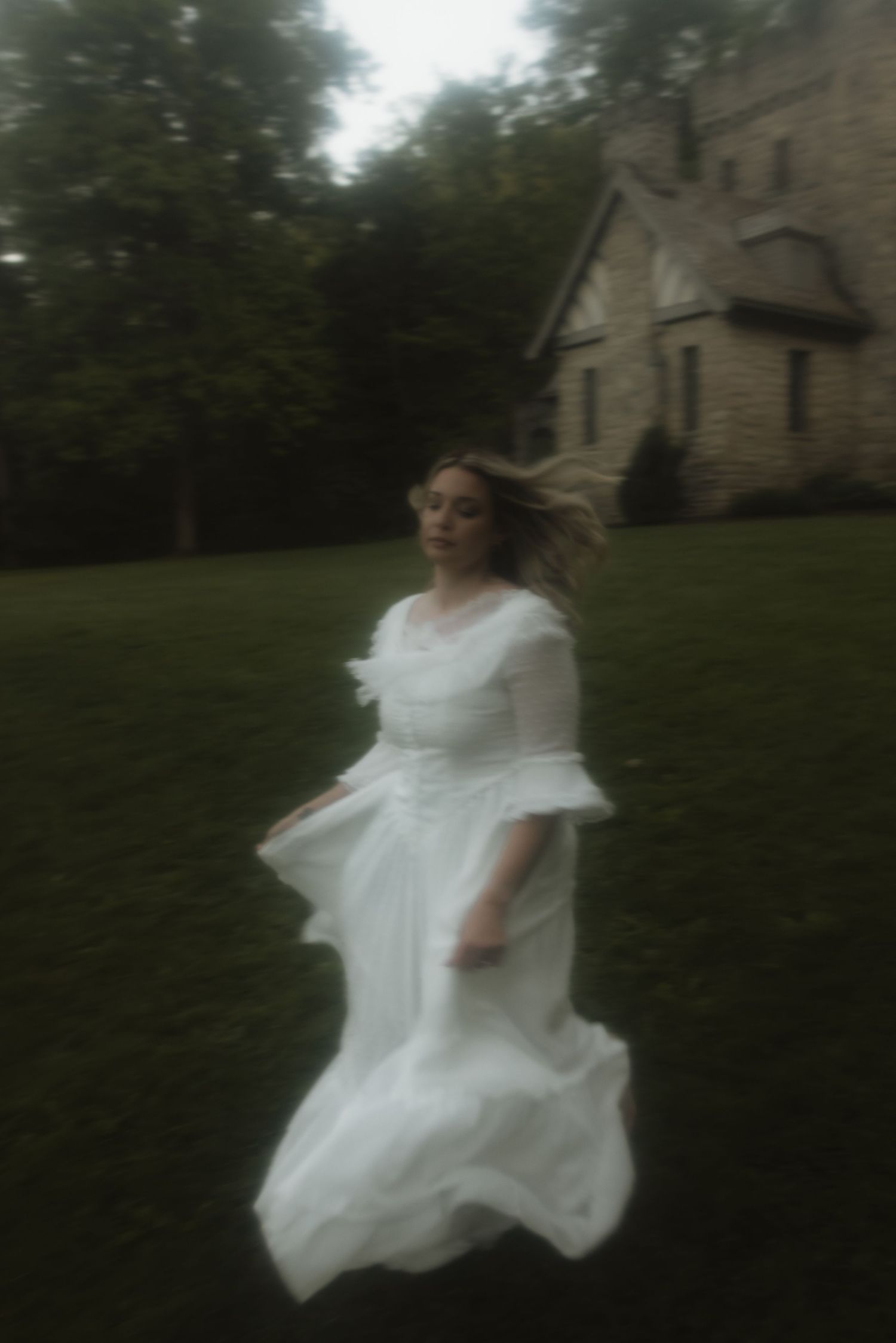 A dreamy blurred photograph shows a figure in a flowing white dress standing in front of a Victorian house on a grassy lawn.