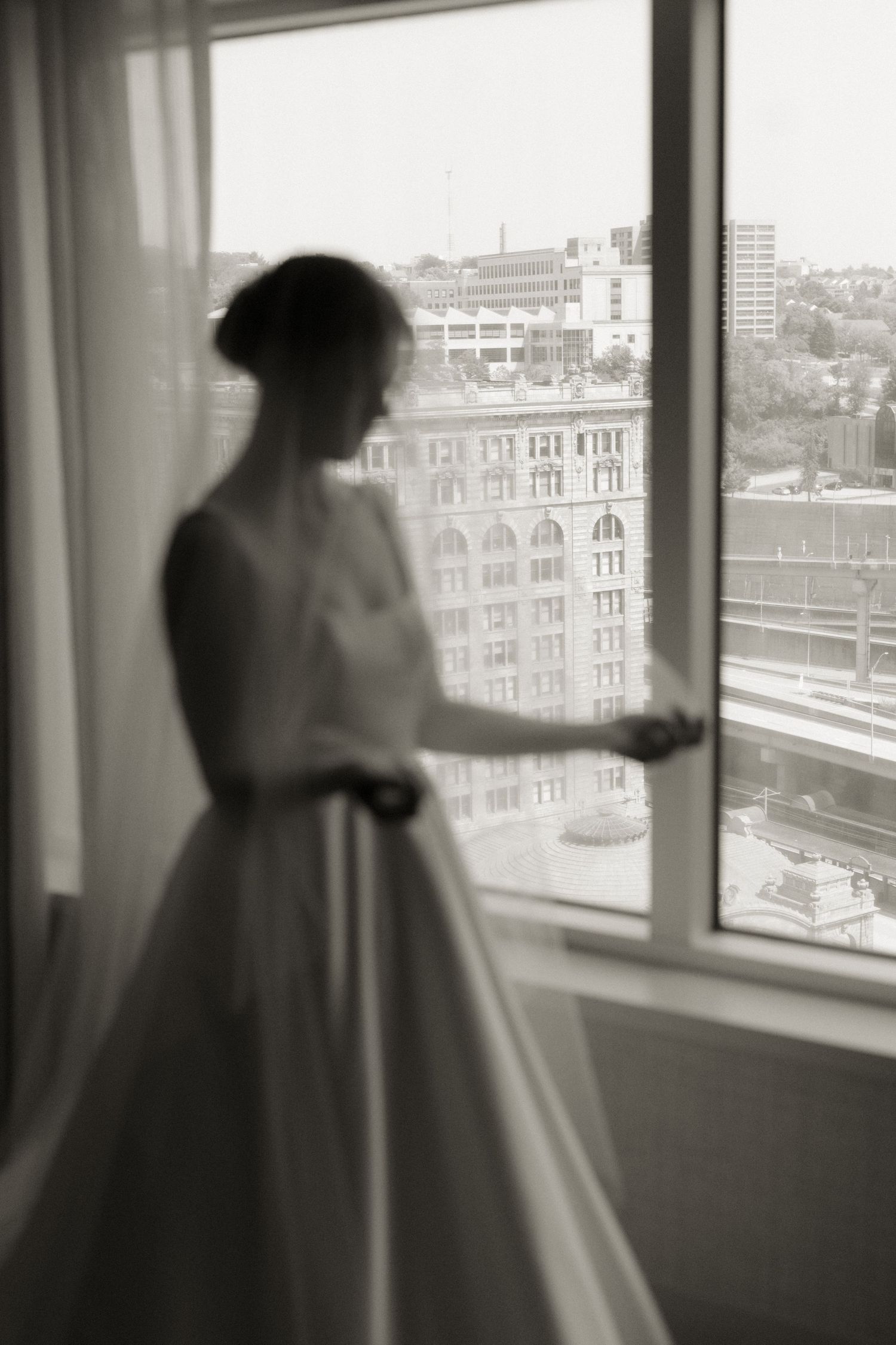 Silhouette of someone in a wedding gown looking out a high-rise window at city buildings.