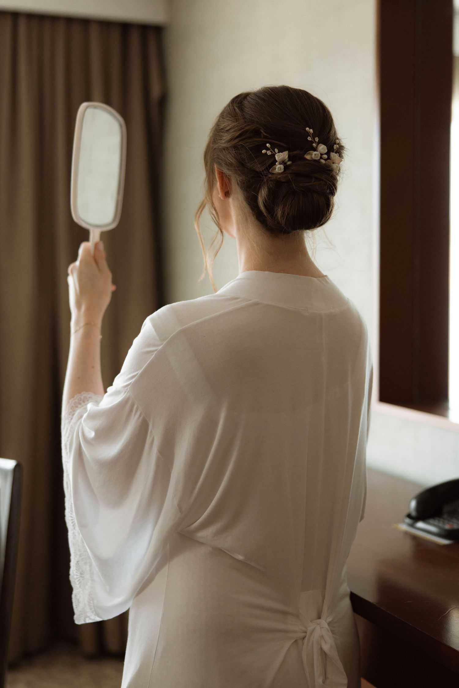 A person in a white robe holds up a hand mirror while wearing an elegant updo hairstyle adorned with delicate accessories.