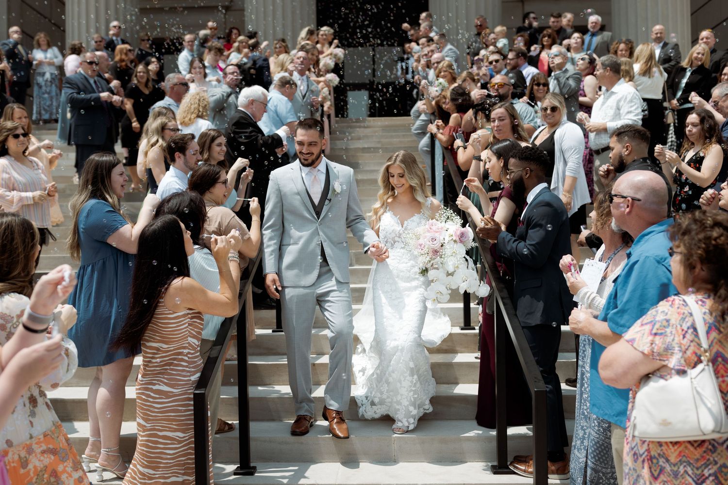 Newlyweds walk down church steps through crowd of wedding guests taking photos in summer.
