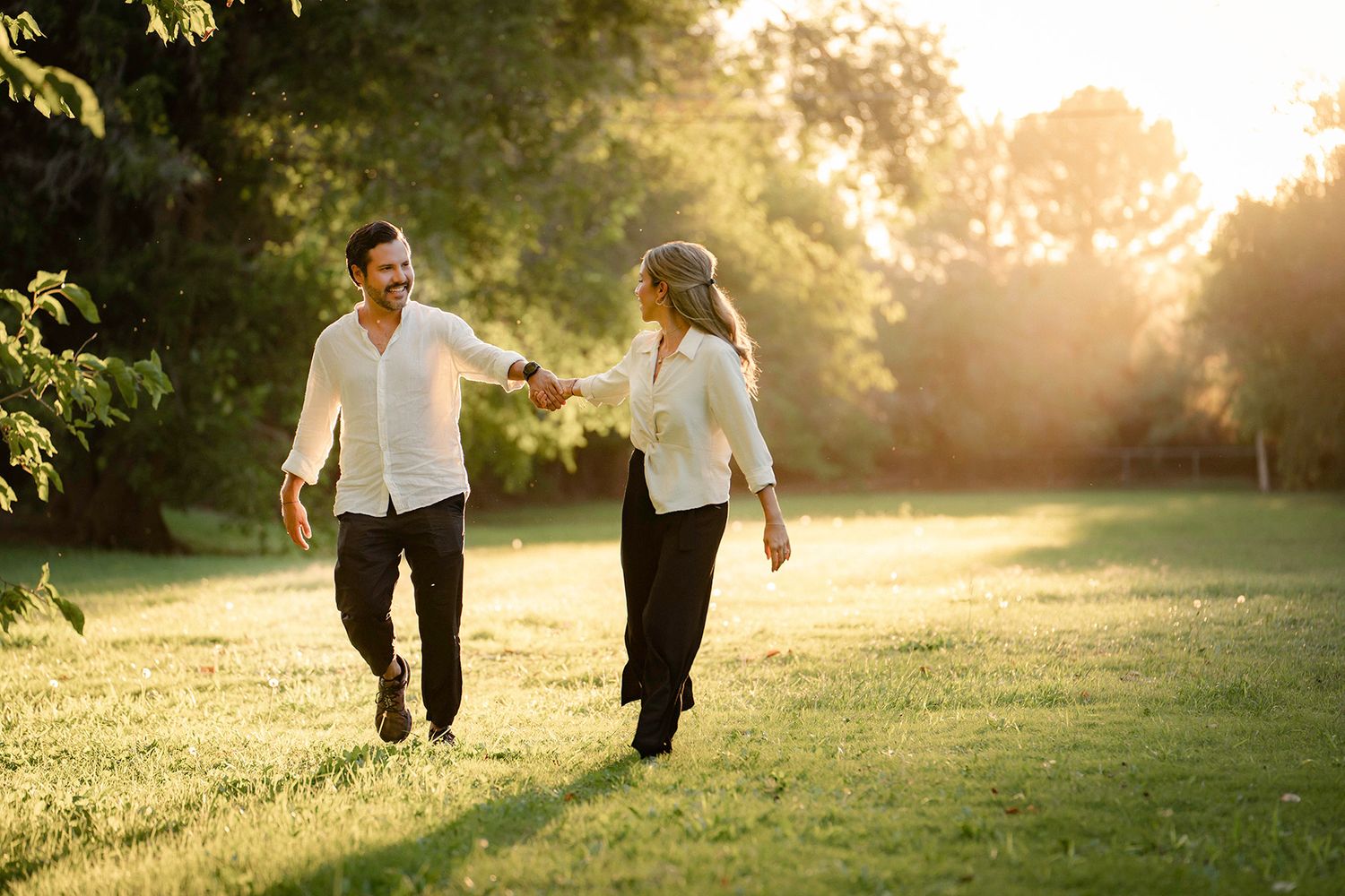 A couple holds hands while walking through a sun-drenched park at sunset.