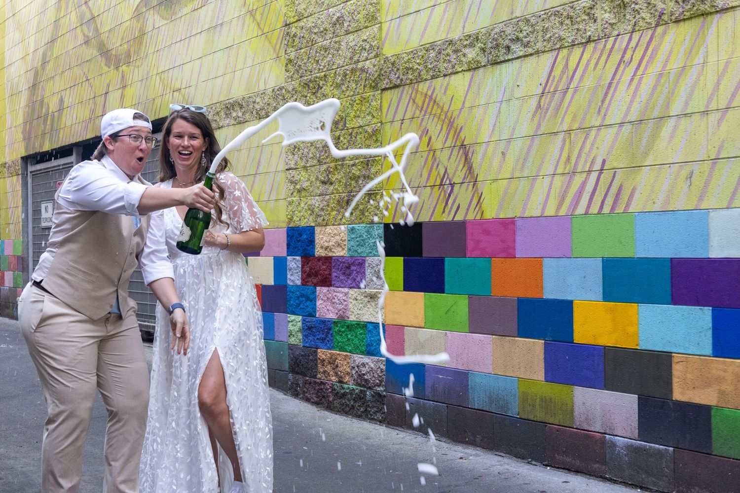 Couple poses playfully in front of a colorful geometric mural wall with confetti floating in the air.