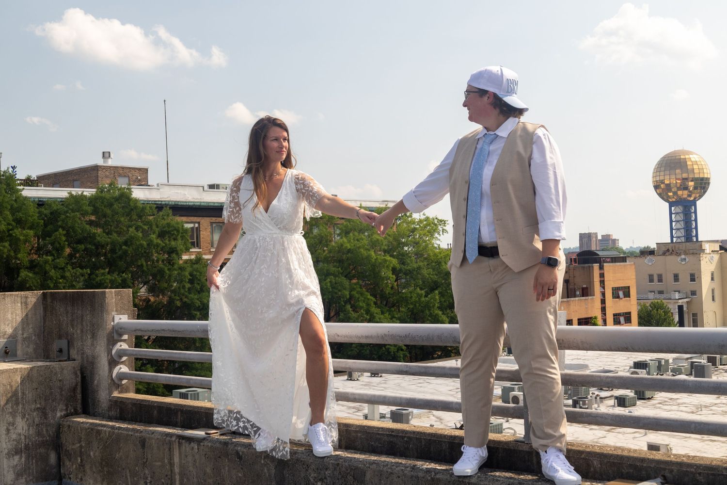 A couple in white wedding attire walk together on a sunny rooftop with city buildings visible in the background.