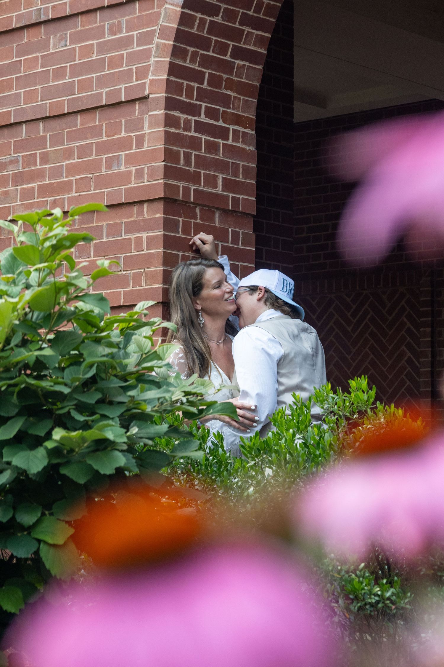 A tender moment captured through pink flowers in front of a brick building with green foliage.