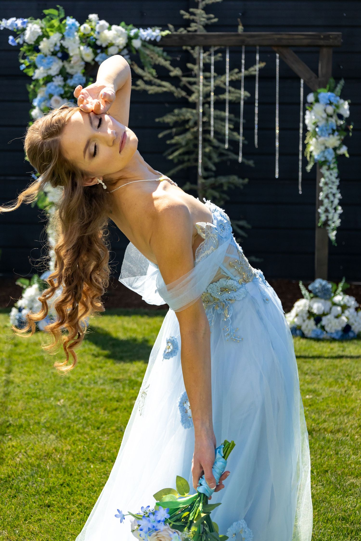 A bride in a white dress poses dramatically with flowers under a floral arch in a garden setting.