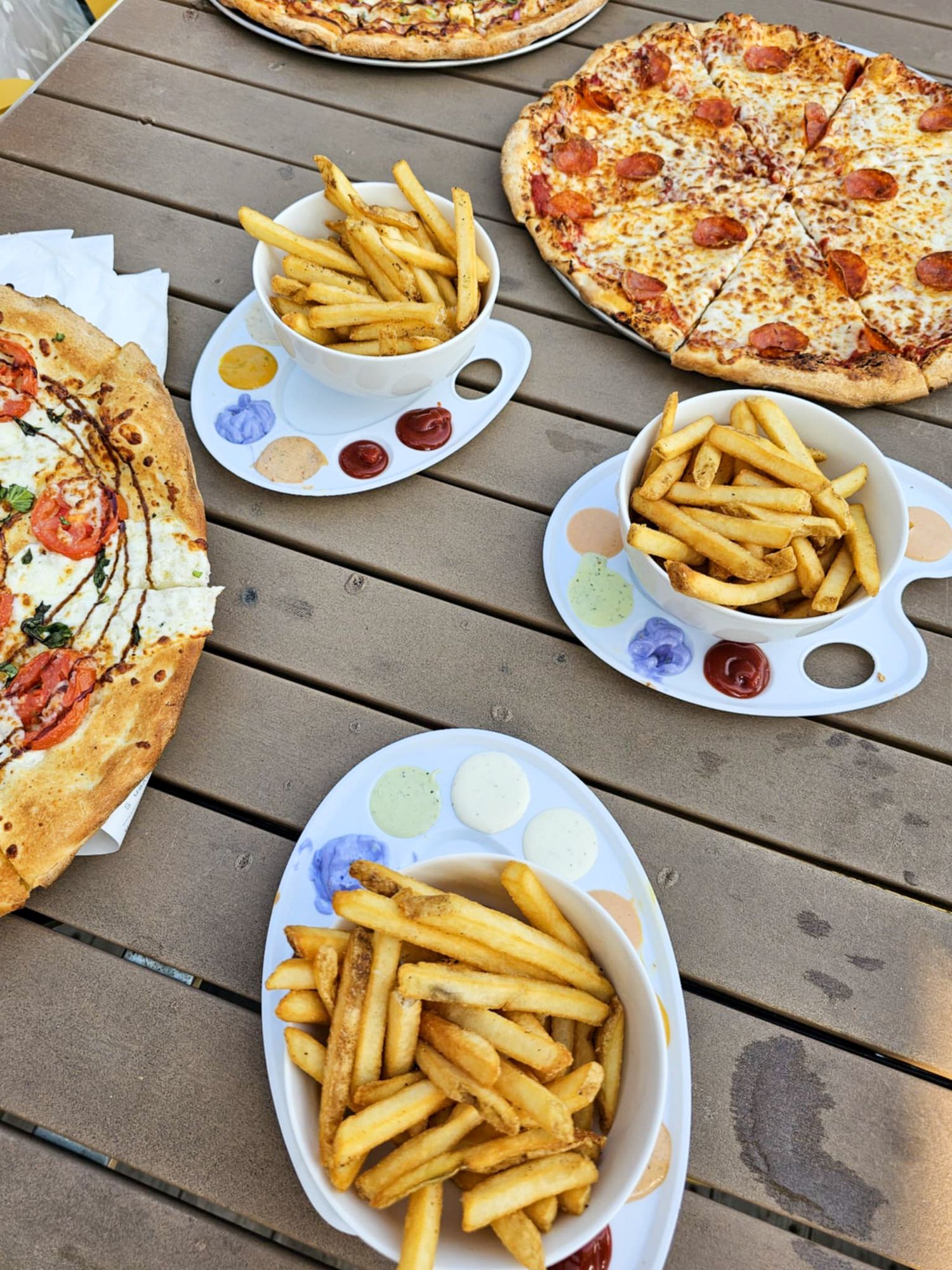 Overhead view of pizzas and french fries served on white plates with assorted dipping sauces on a wooden outdoor table.