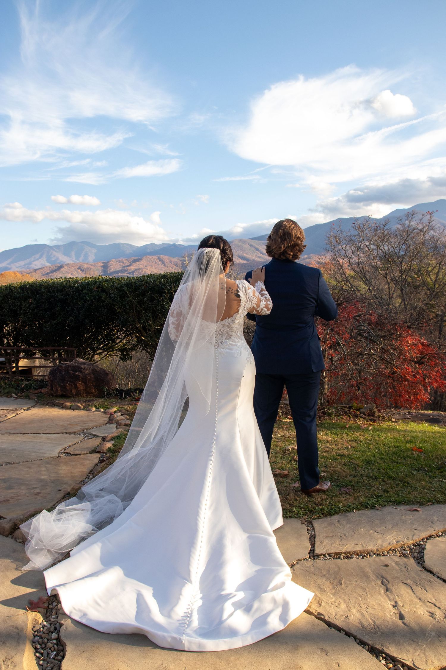 Newlyweds admire mountain views from stone patio with bride's long train and veil flowing behind.