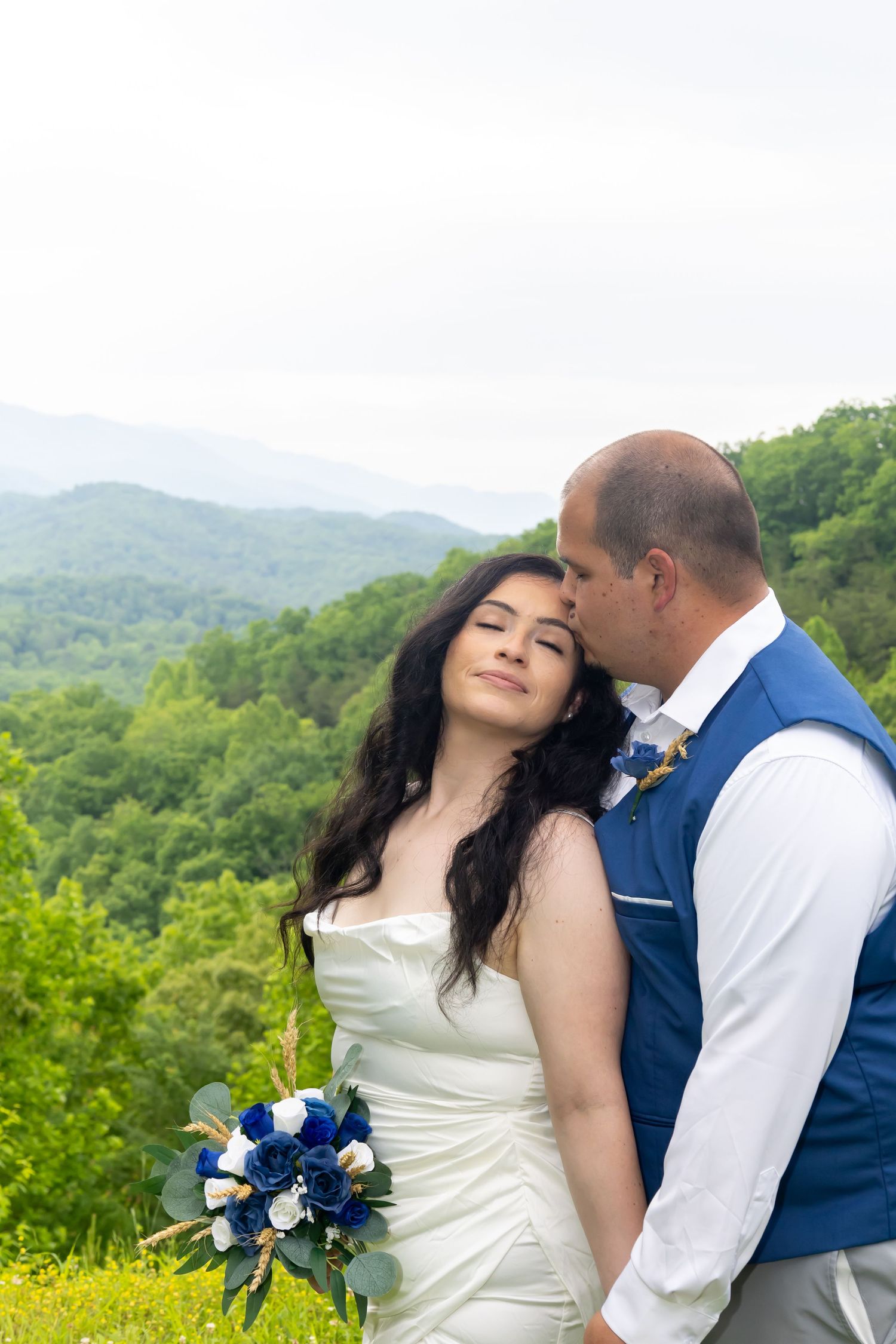 Romantic wedding portrait with mountain views as couple embraces holding blue and white bouquet.