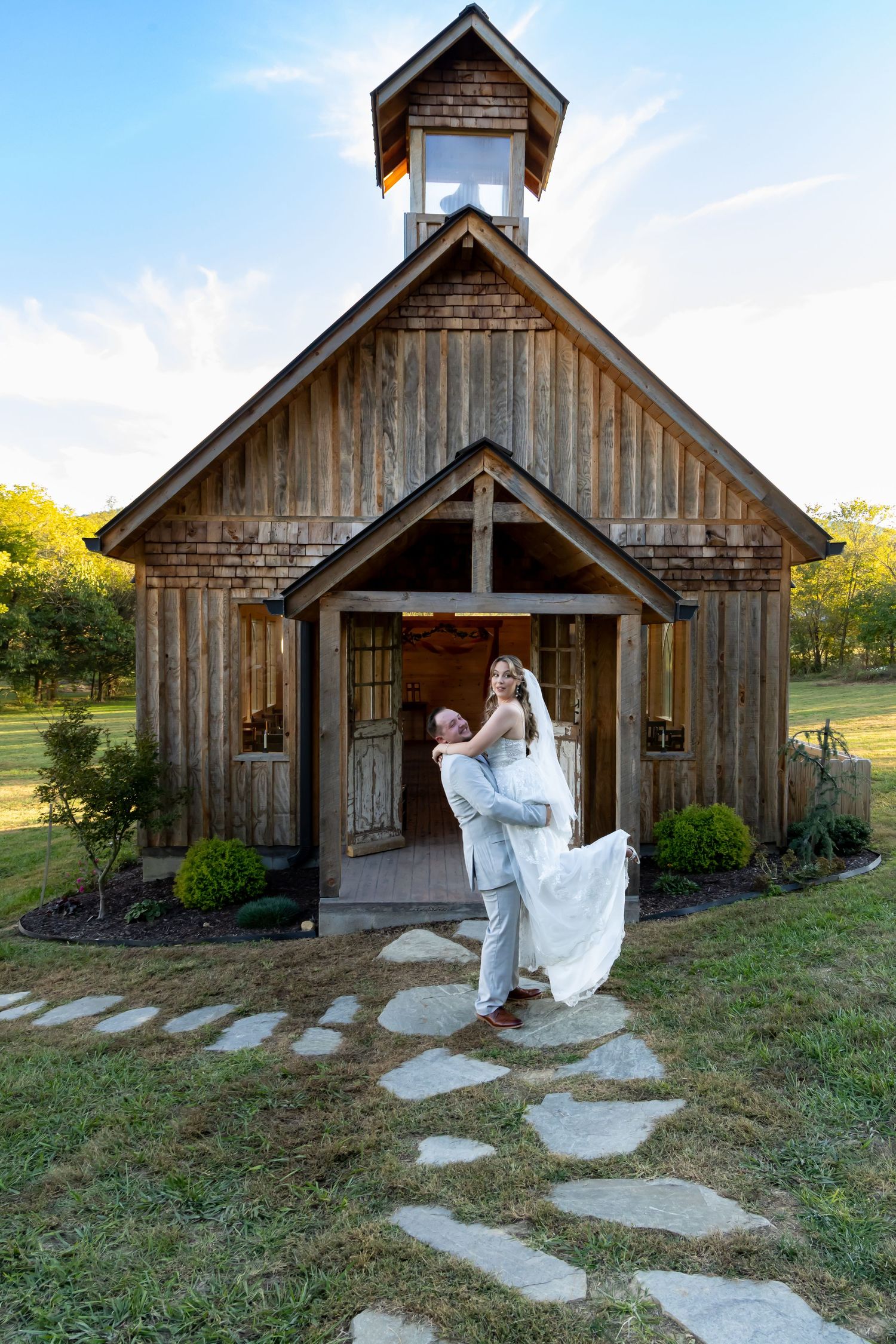 Rustic chapel wedding scene with couple posed on stone pathway leading to wooden church entrance.