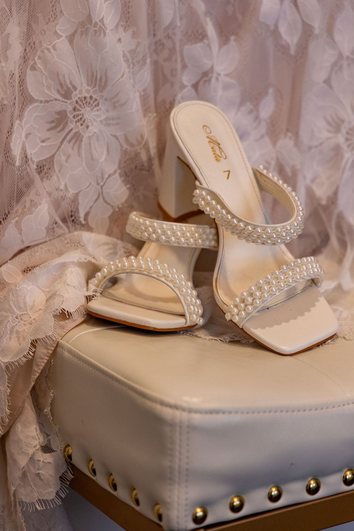 Pearl-embellished cream wedding heels displayed on a studded white leather ottoman.