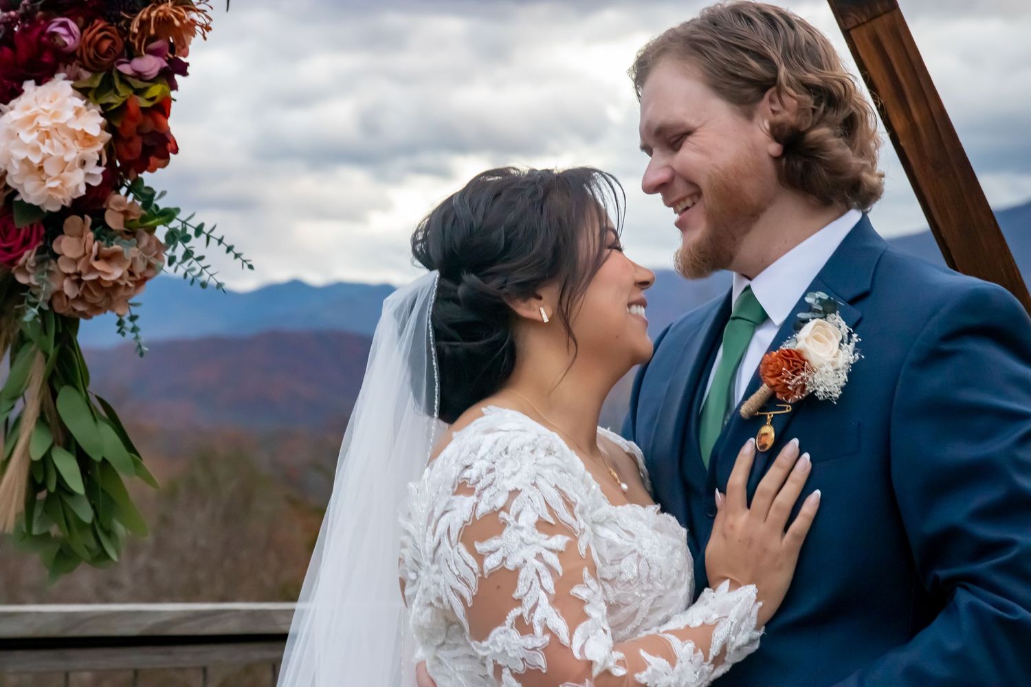 Wedding couple shares intimate moment under floral arch with mountain landscape in background.