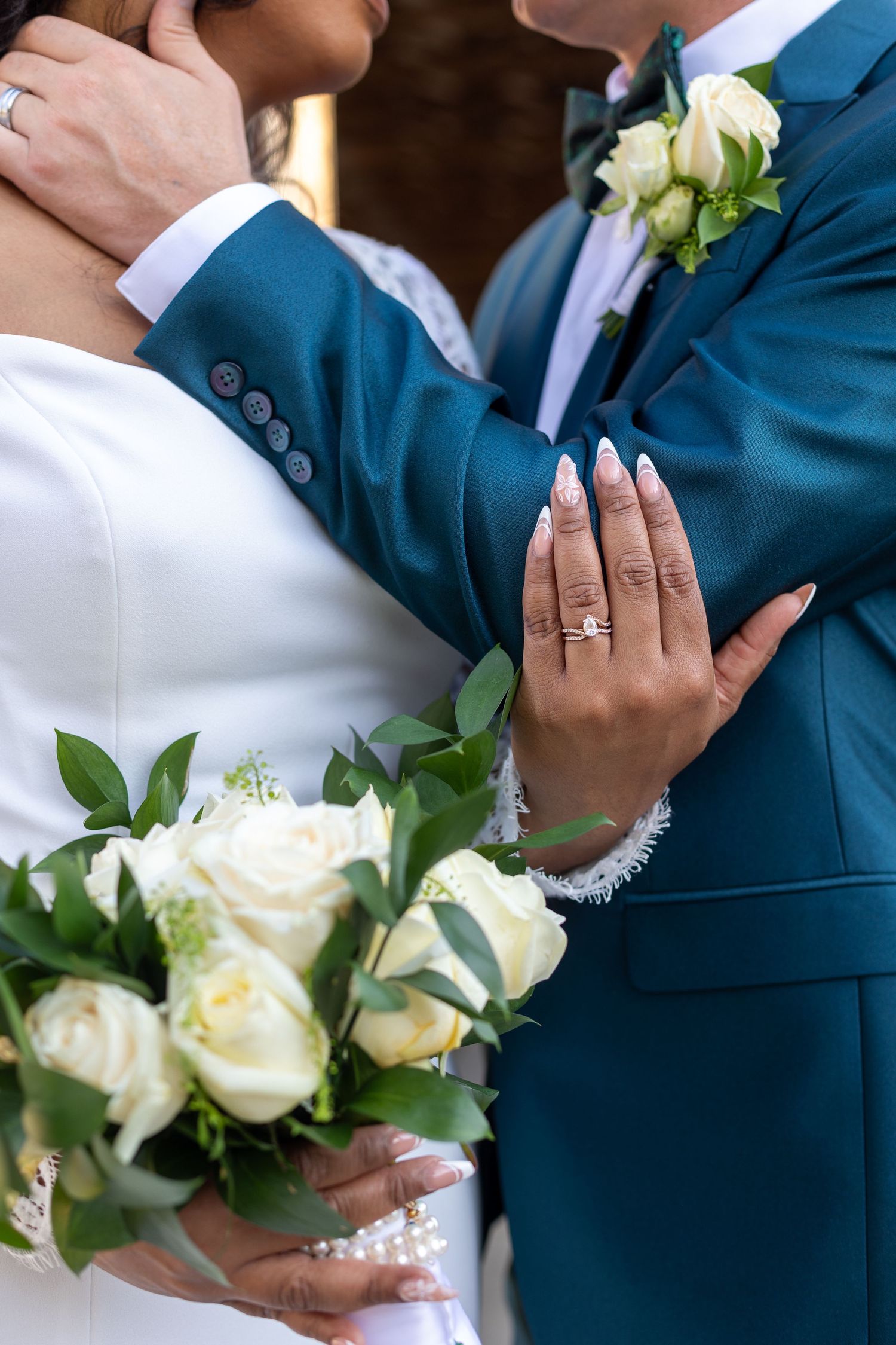 Close-up of wedding rings and white rose bouquet with hands embracing in teal blue suit.