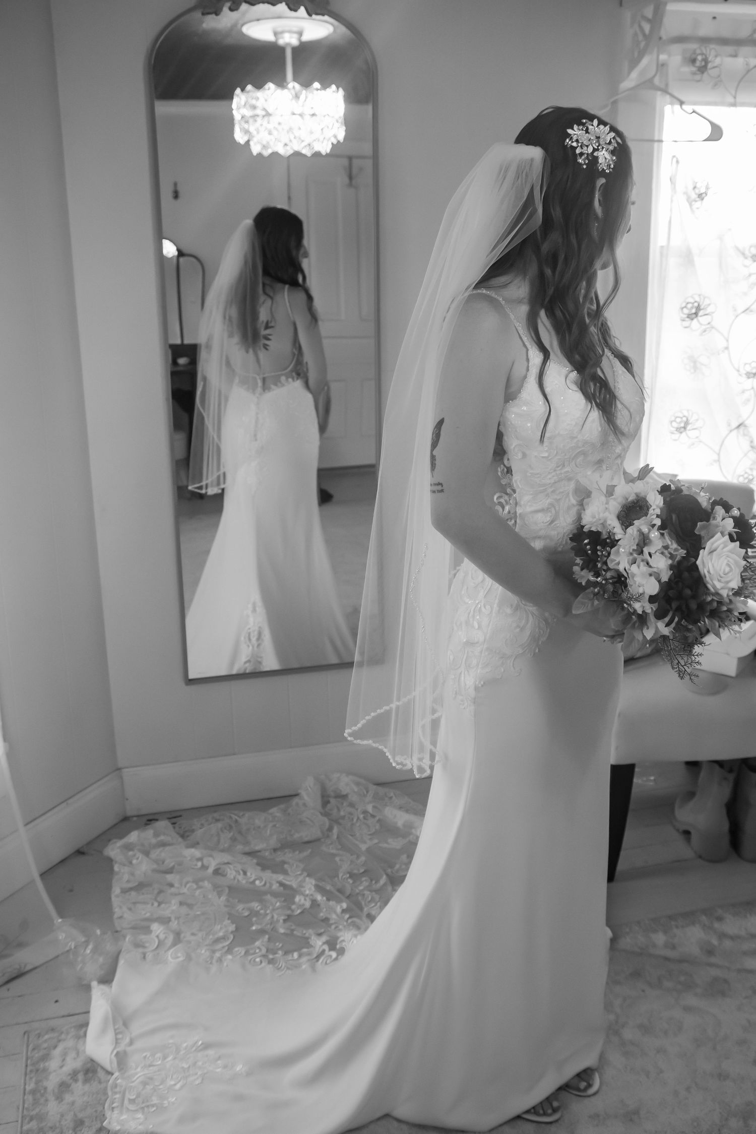 Elegant bride in white gown and veil looks at her reflection in a full-length mirror next to a crystal chandelier.