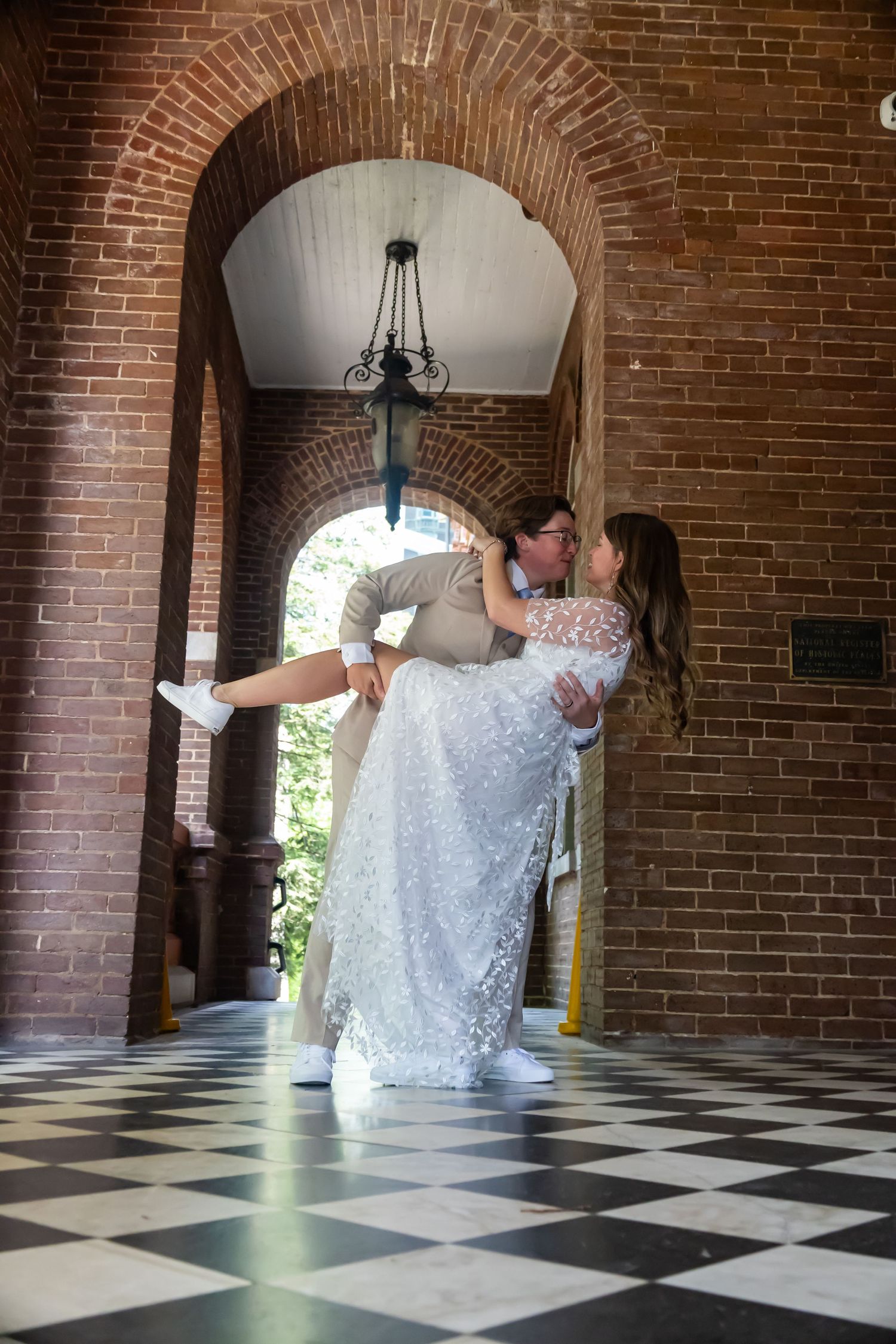 Two people share a romantic dance pose on a checkered floor beneath a brick archway with hanging chandelier.