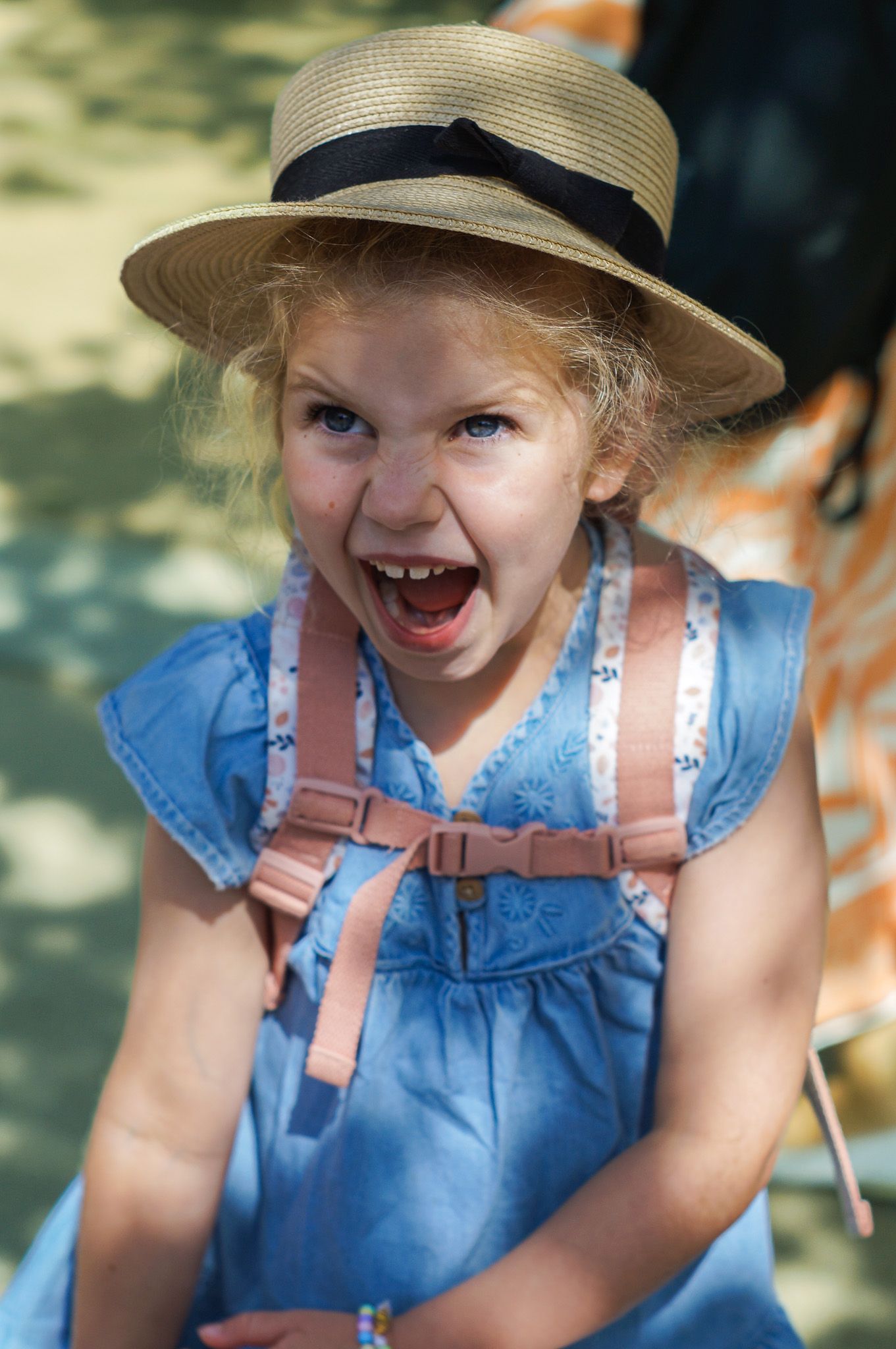 Excited child wearing a straw sun hat and blue dress with pink overalls laughs outdoors on a sunny day.