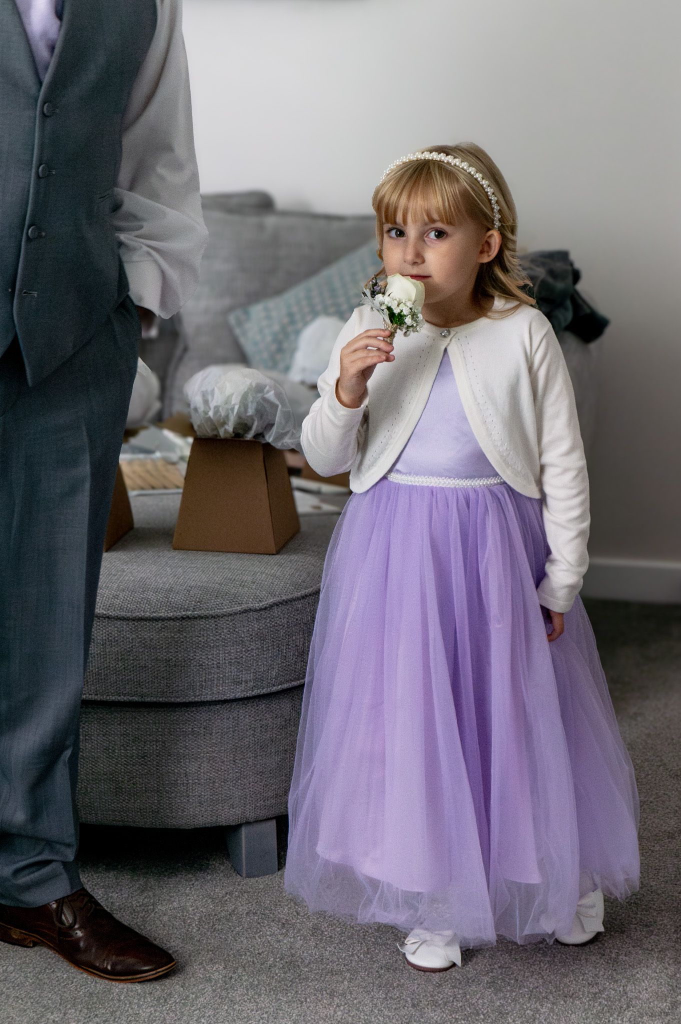 A small child in a lavender tulle dress and white cardigan stands by a gray sofa while eating a snack.