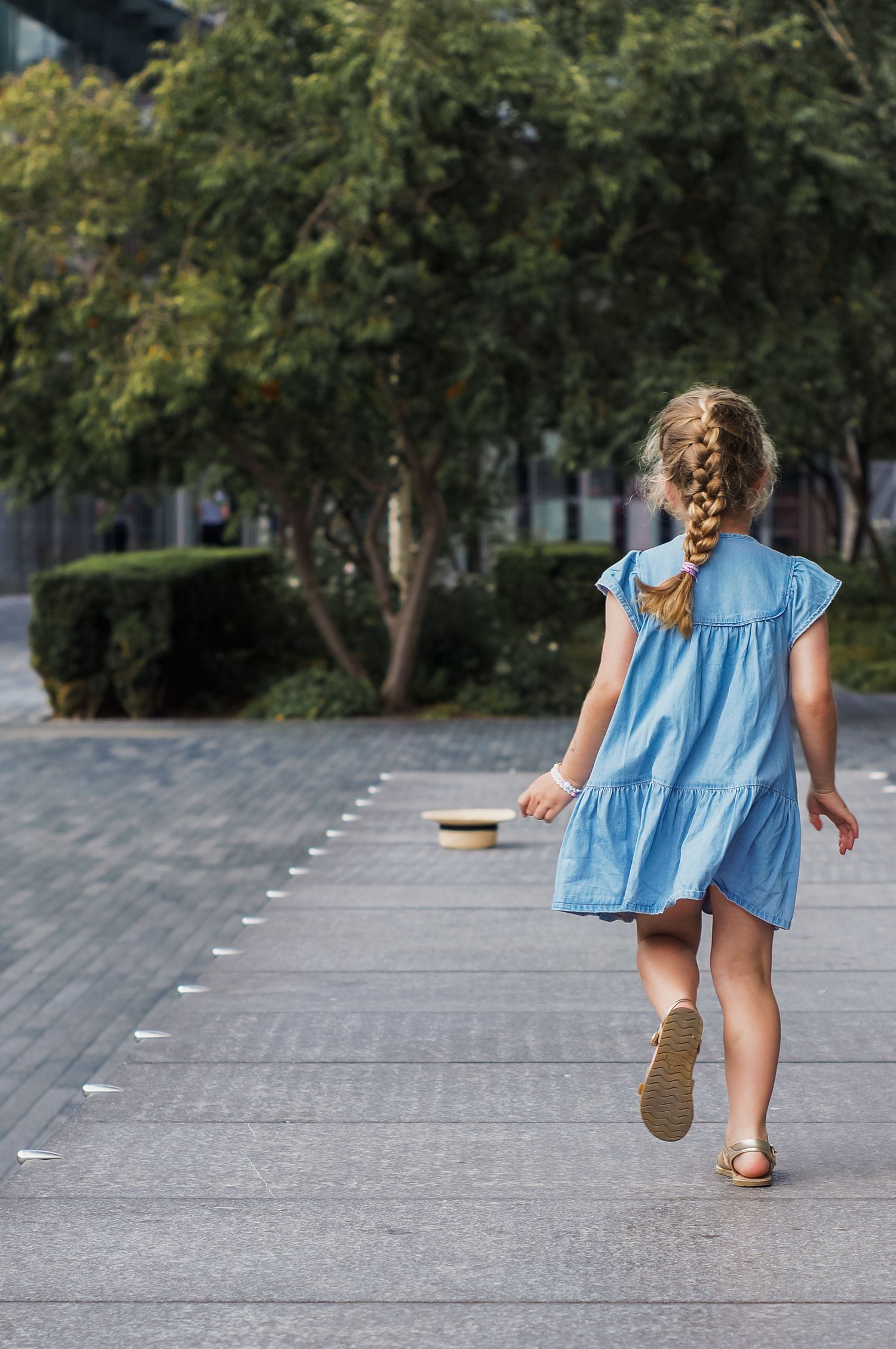 A figure in a light blue tiered dress with braided hair walks down a paved path on a summer day.