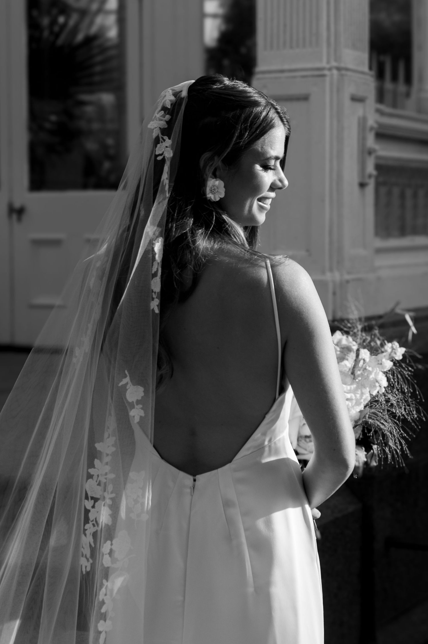Black and white portrait of a bride in a backless wedding dress and veil smiling while looking to the side.