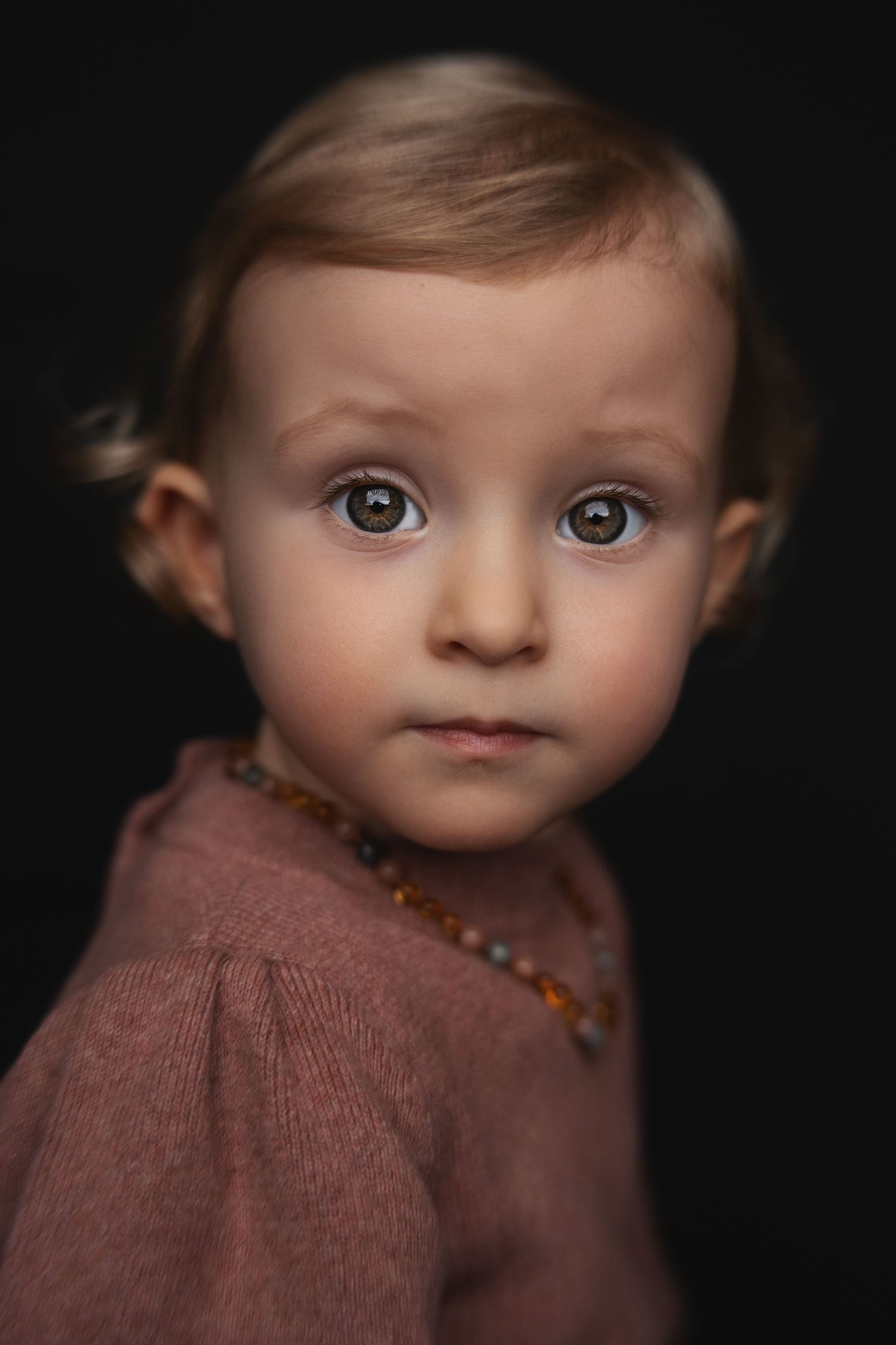 Close-up portrait of a young child with striking blue eyes wearing a mauve sweater against a dark background.