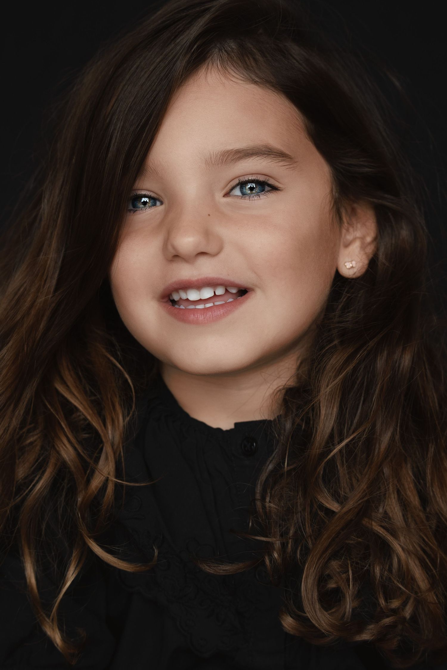 A close-up portrait of a child with long brown wavy hair wearing a black top and smiling warmly at the camera.