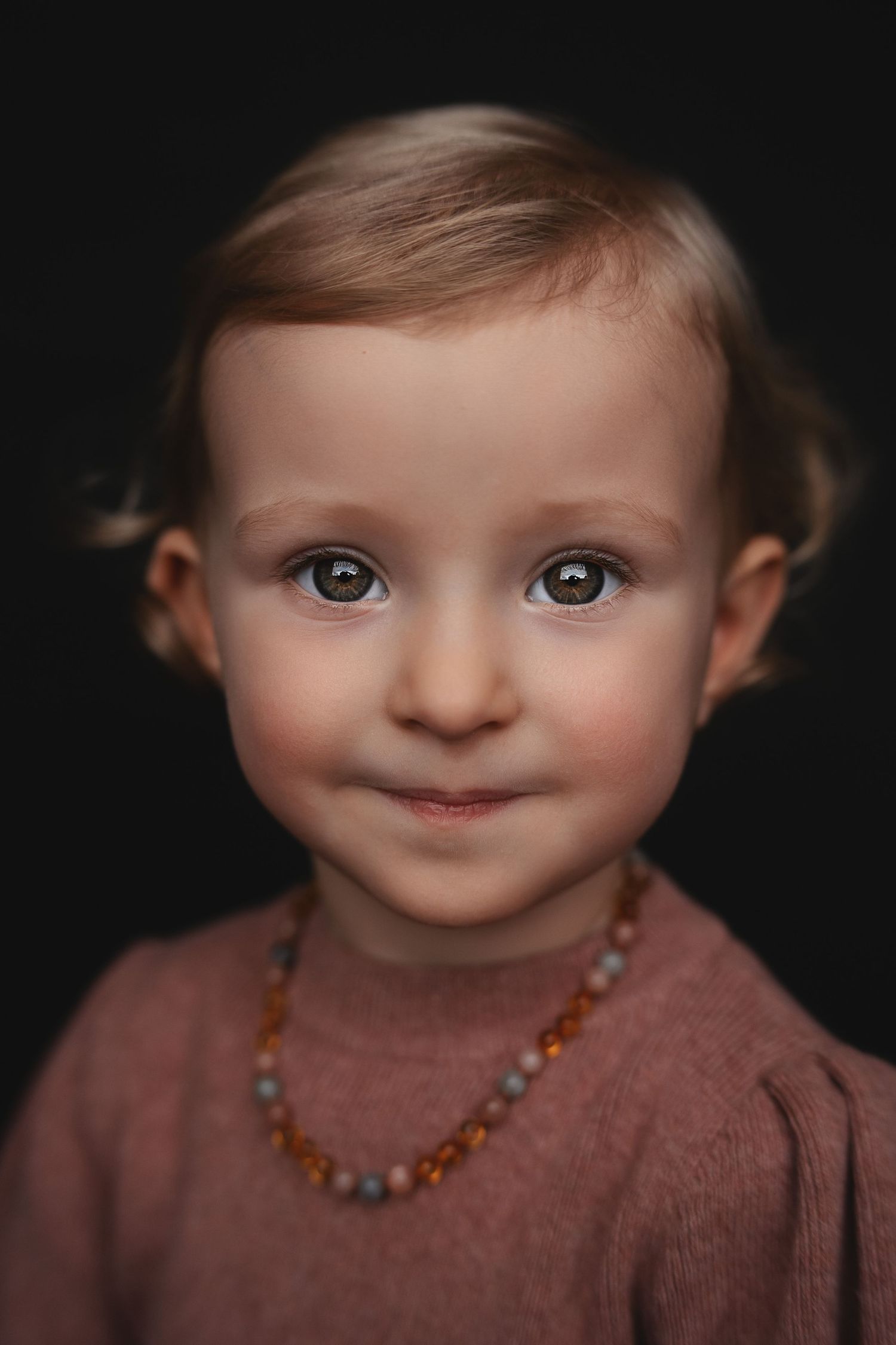 A portrait of a young child in a mauve sweater wearing amber beads against a dark background.