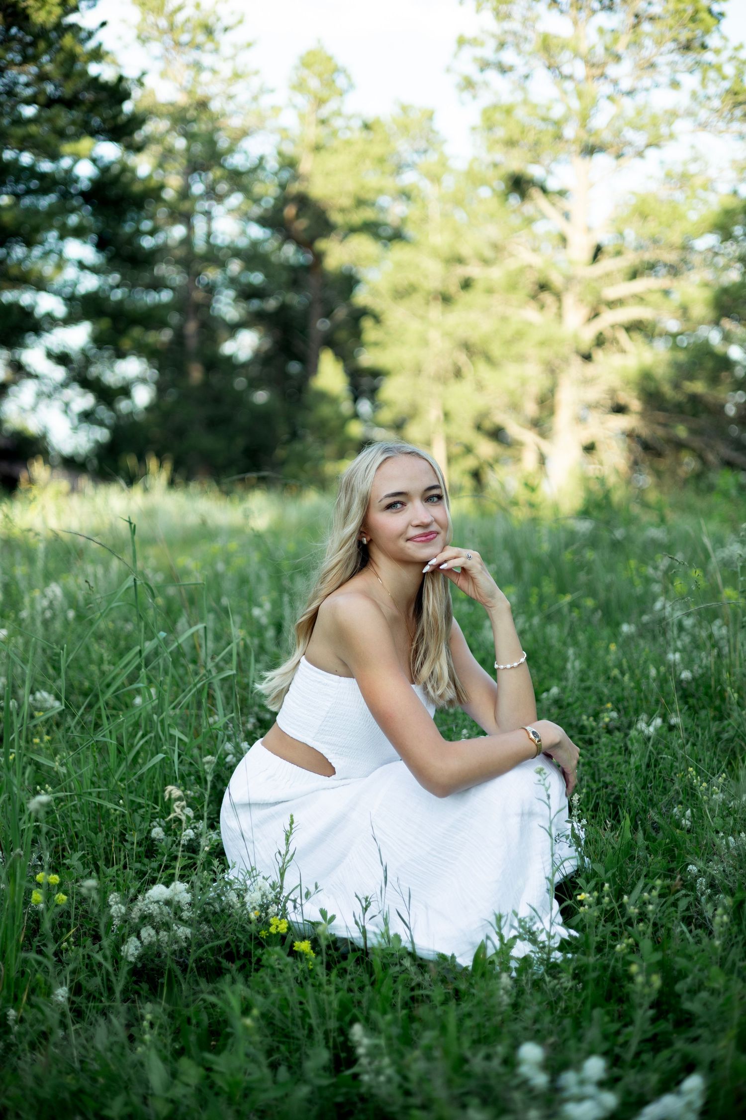 Graceful portrait in a white dress sitting among wildflowers and tall grass with evergreen trees in background.