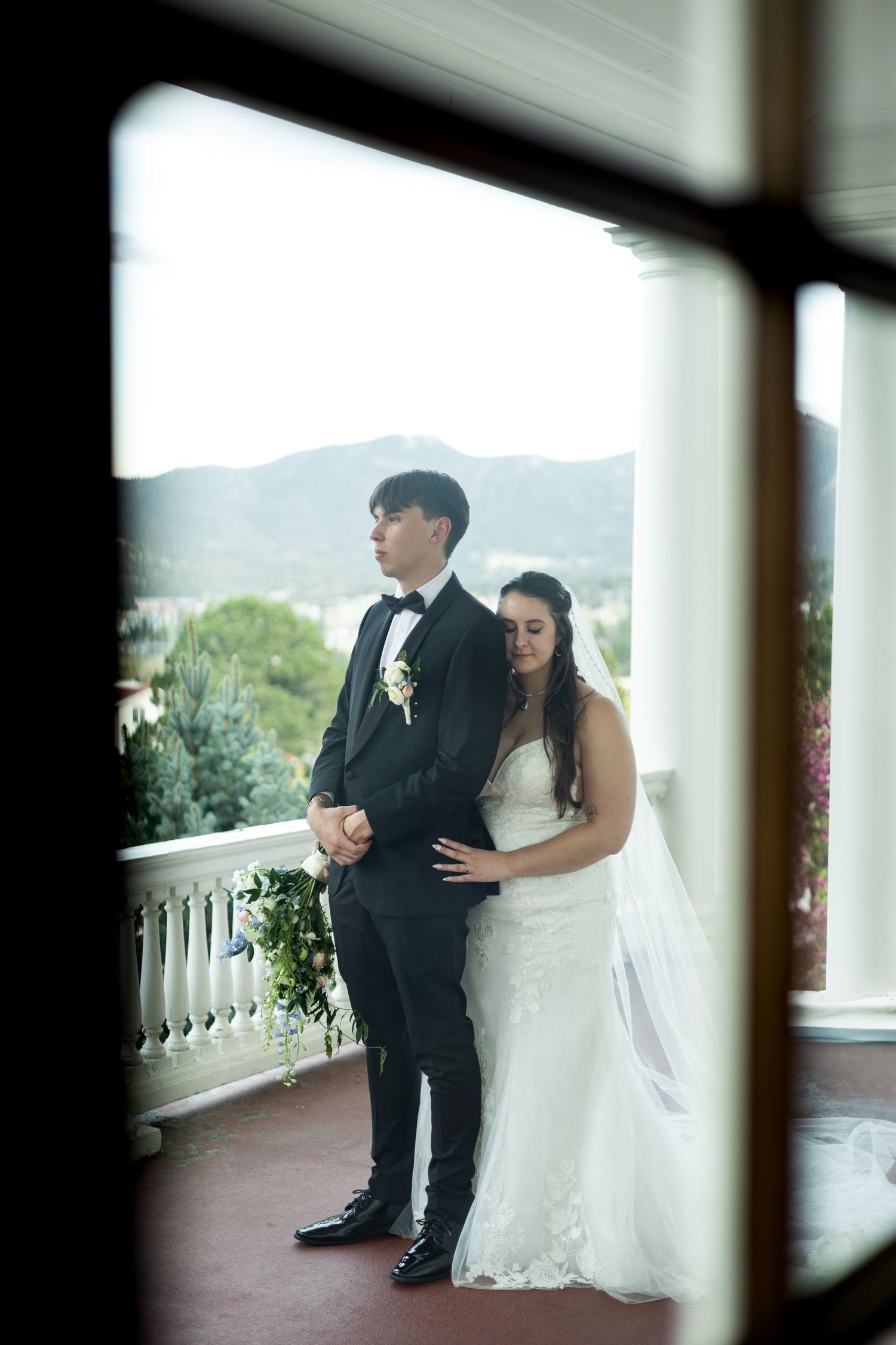 Newlywed couple stands on elegant balcony with mountain vista in background viewed through window frame.