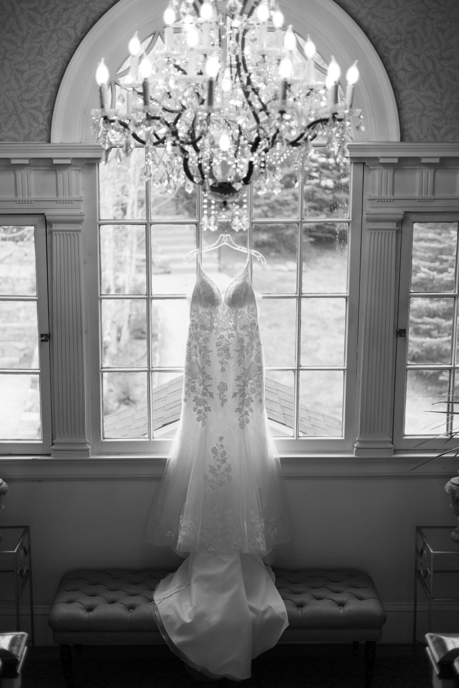 An elegant wedding dress hangs from a crystal chandelier in front of an arched window, captured in black and white.