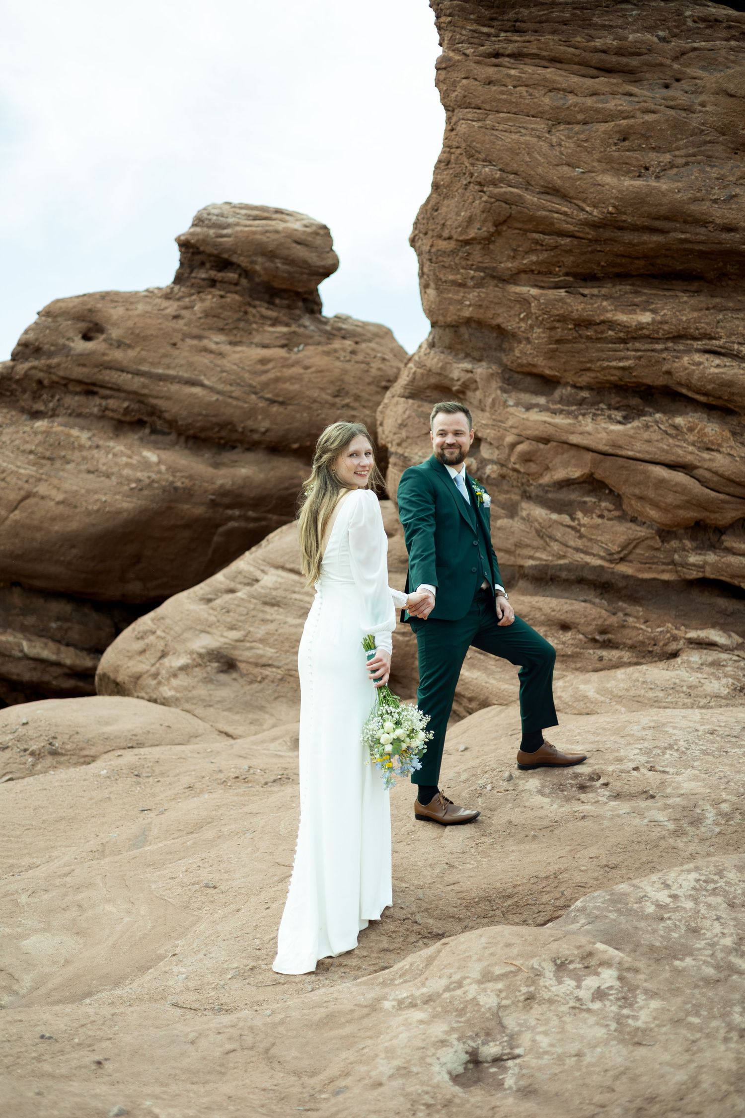 A couple in wedding attire poses together on red rock formations in a desert landscape.