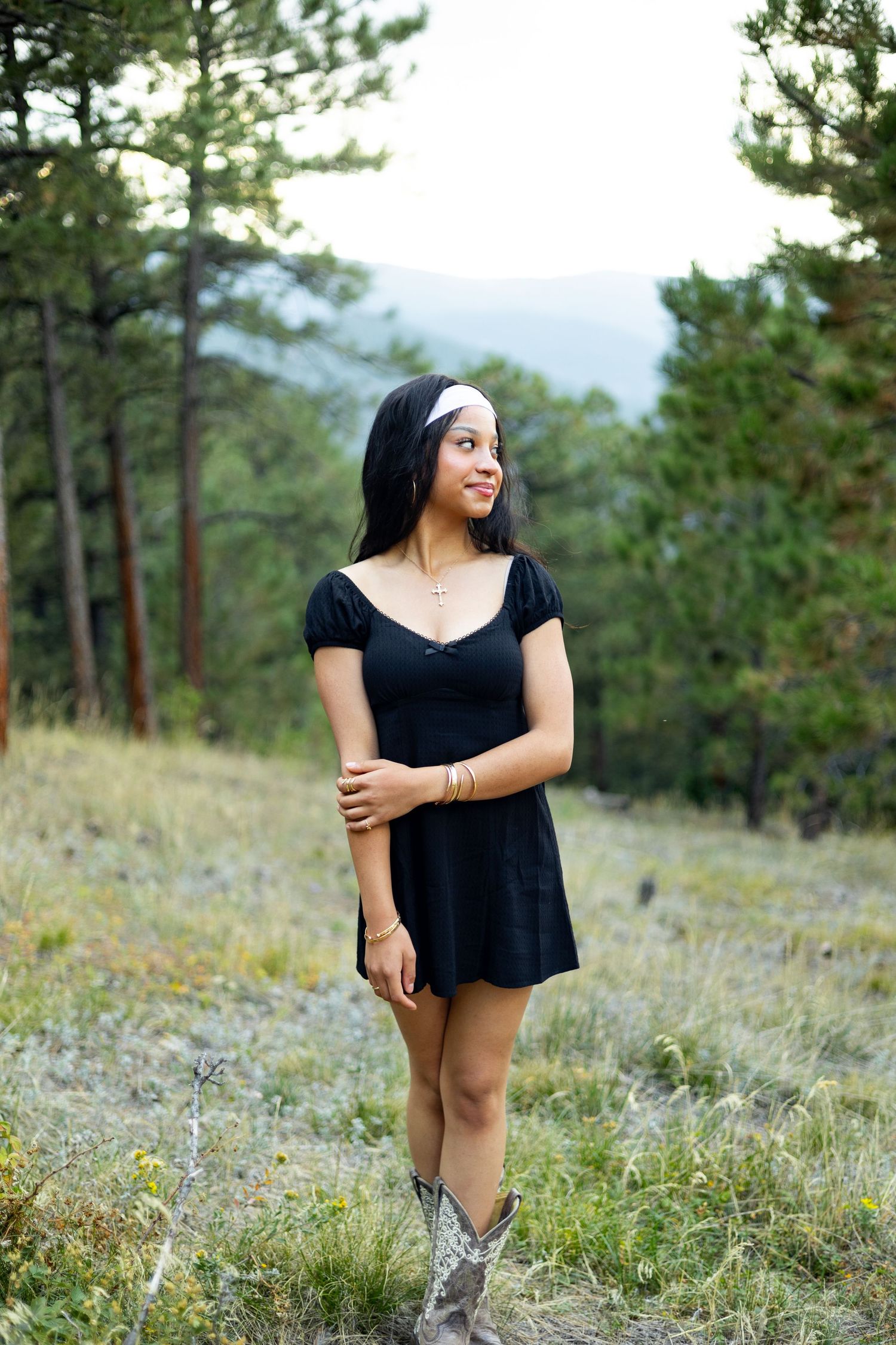 Woman in black dress poses in mountain meadow with pine trees.