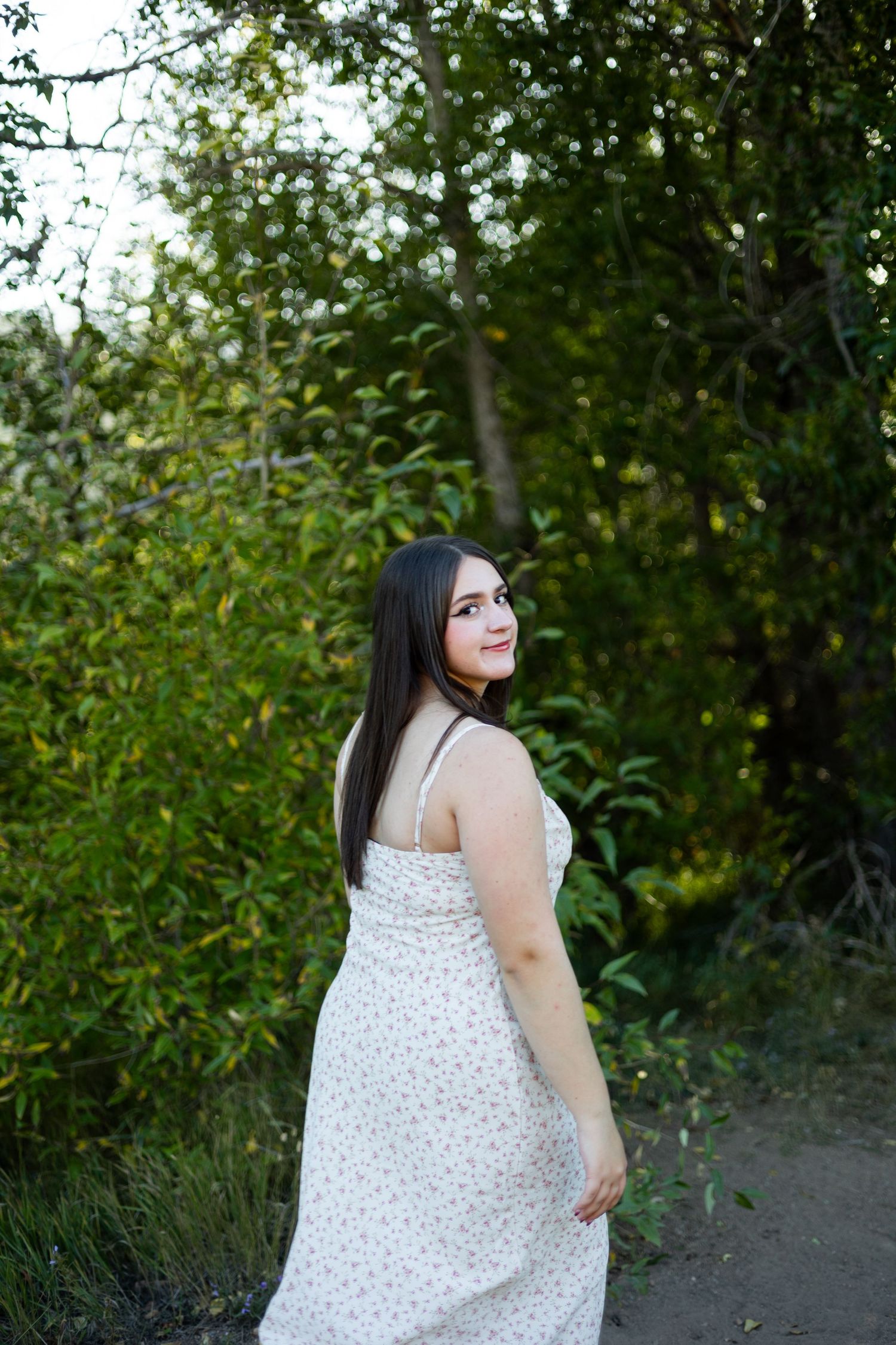Woman in white floral dress poses on nature path with greenery background.