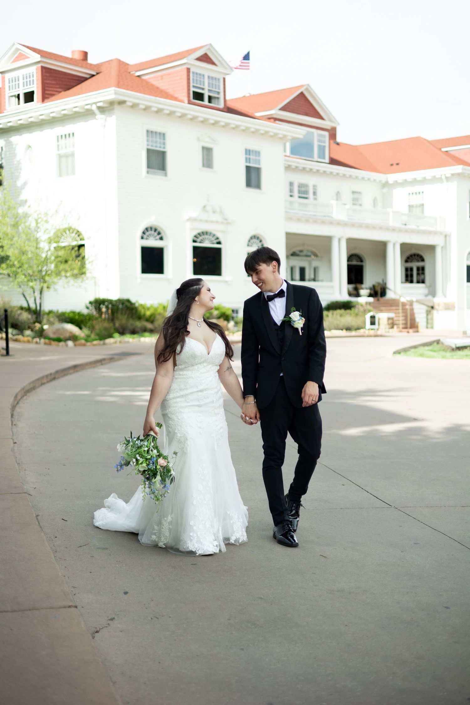 A bride in white lace dress and groom in black tuxedo stand outside a grand colonial-style mansion.