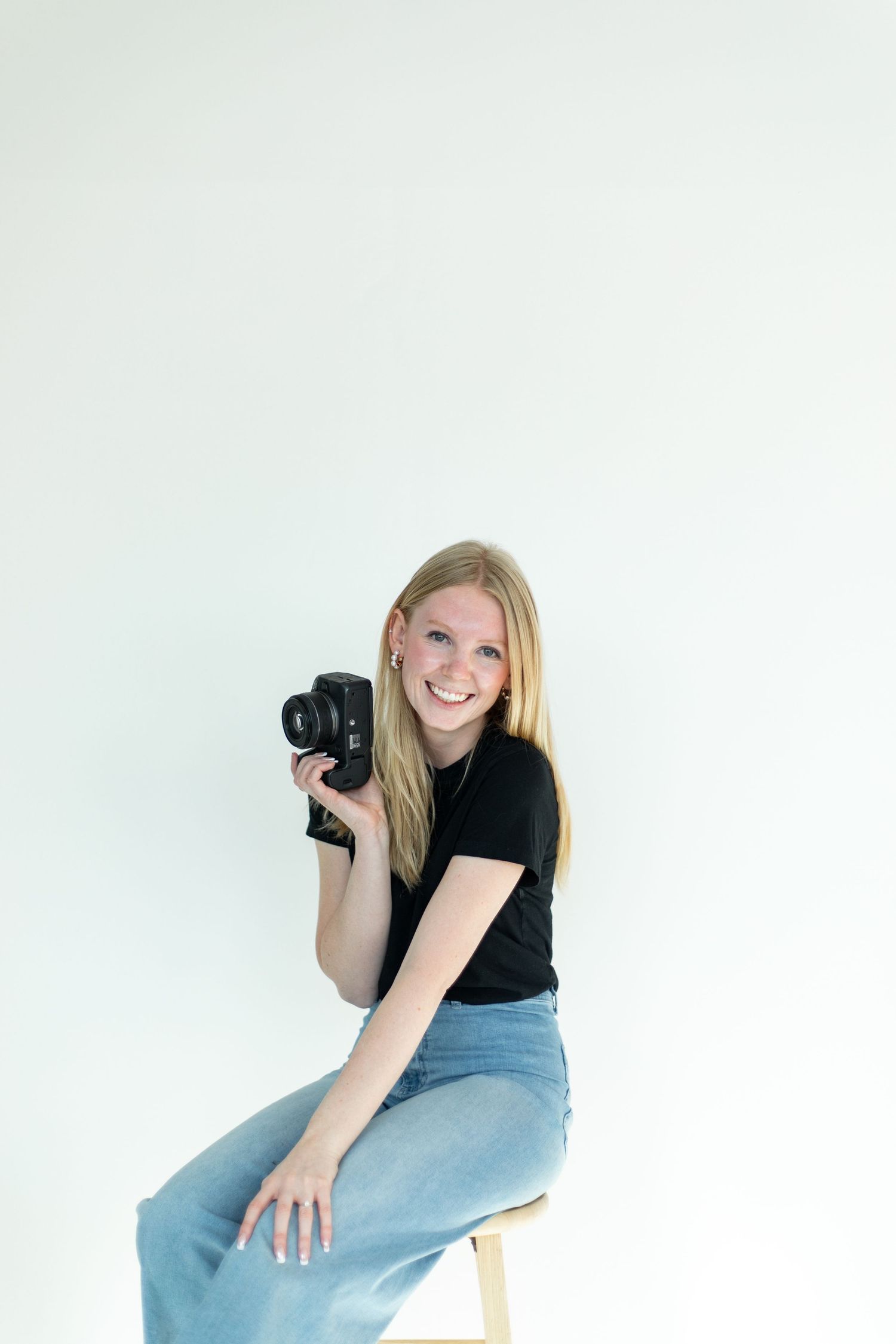 A photographer in casual attire holds a camera while sitting on a stool against a white background.