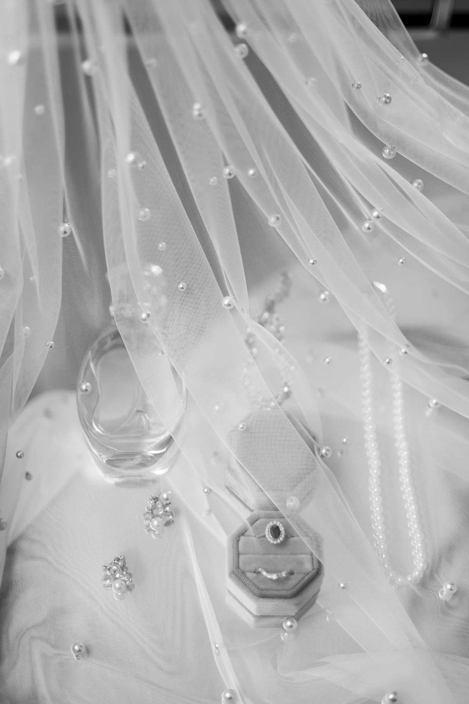 Elegant wedding veil with delicate beading and rhinestone details draped over white fabric in black and white photograph.