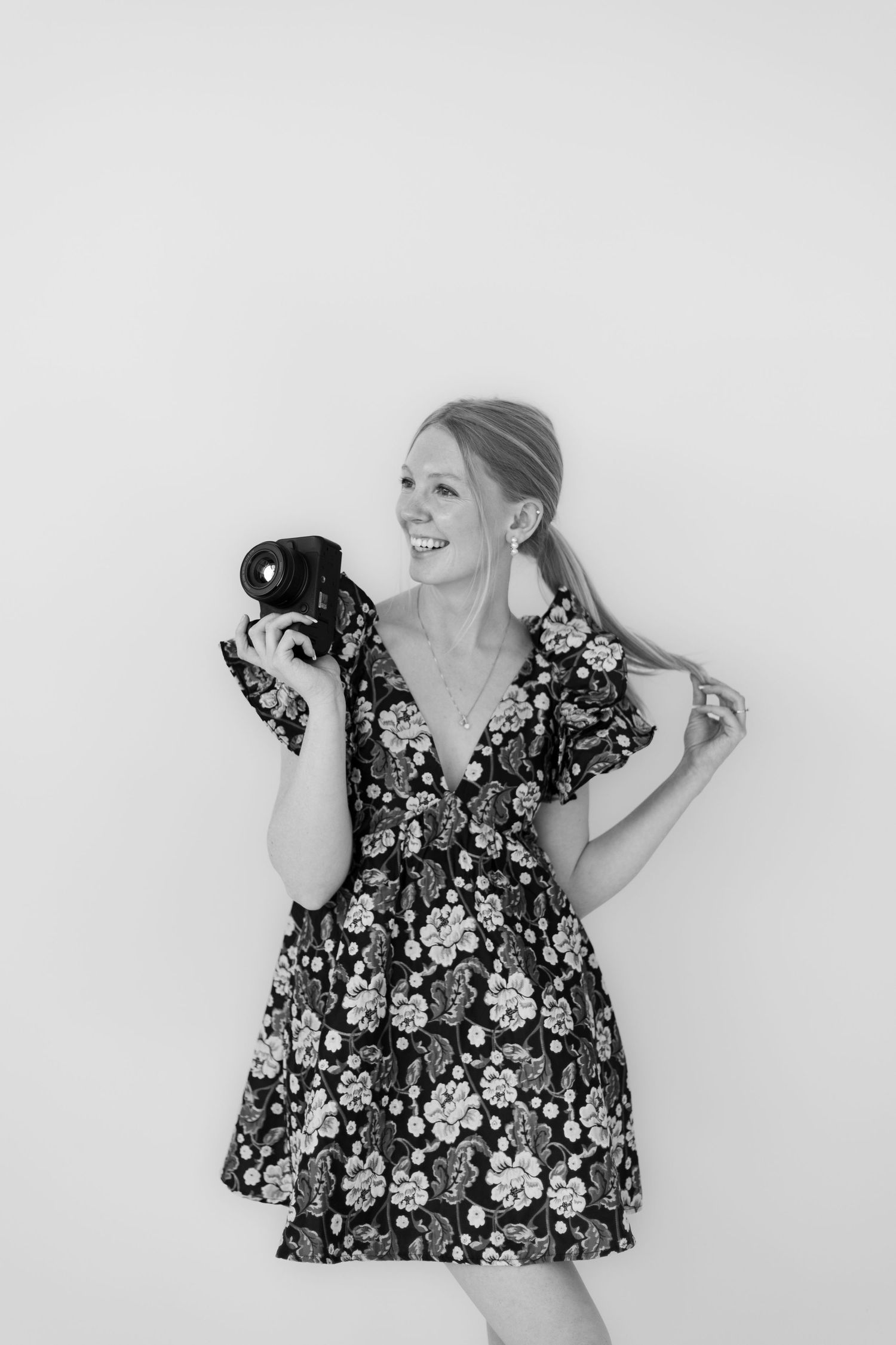 Black and white portrait of photographer in floral dress holding camera.