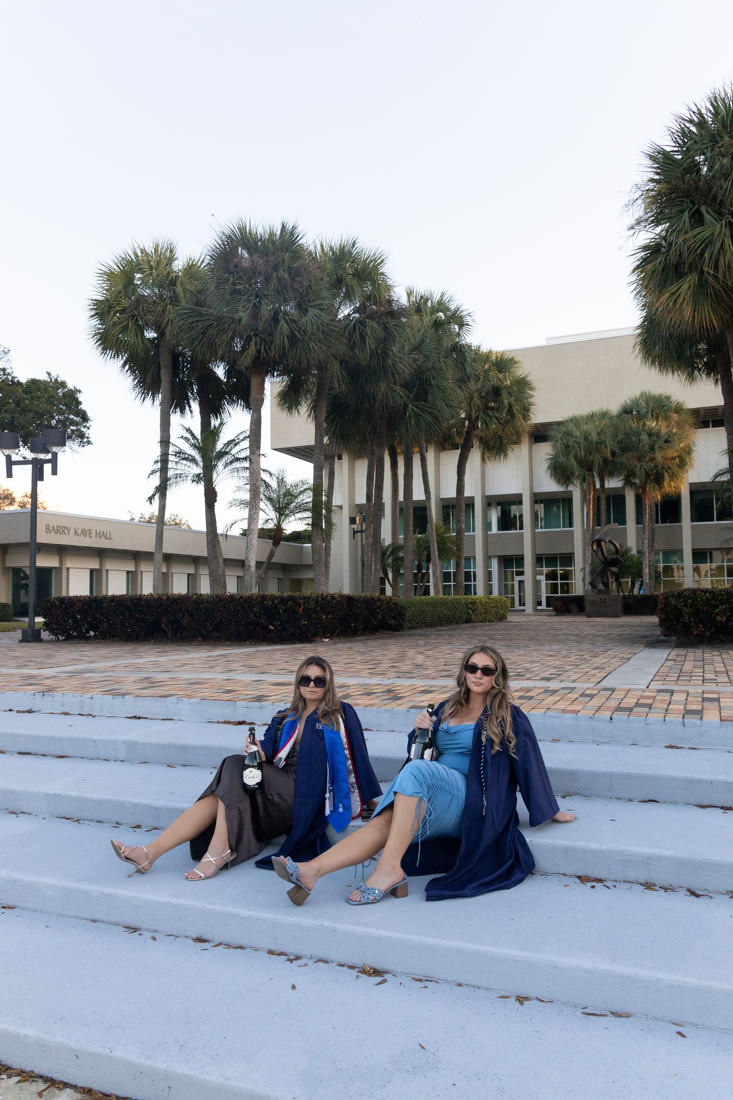Two friends relaxing on concrete steps in front of a modern building surrounded by palm trees.