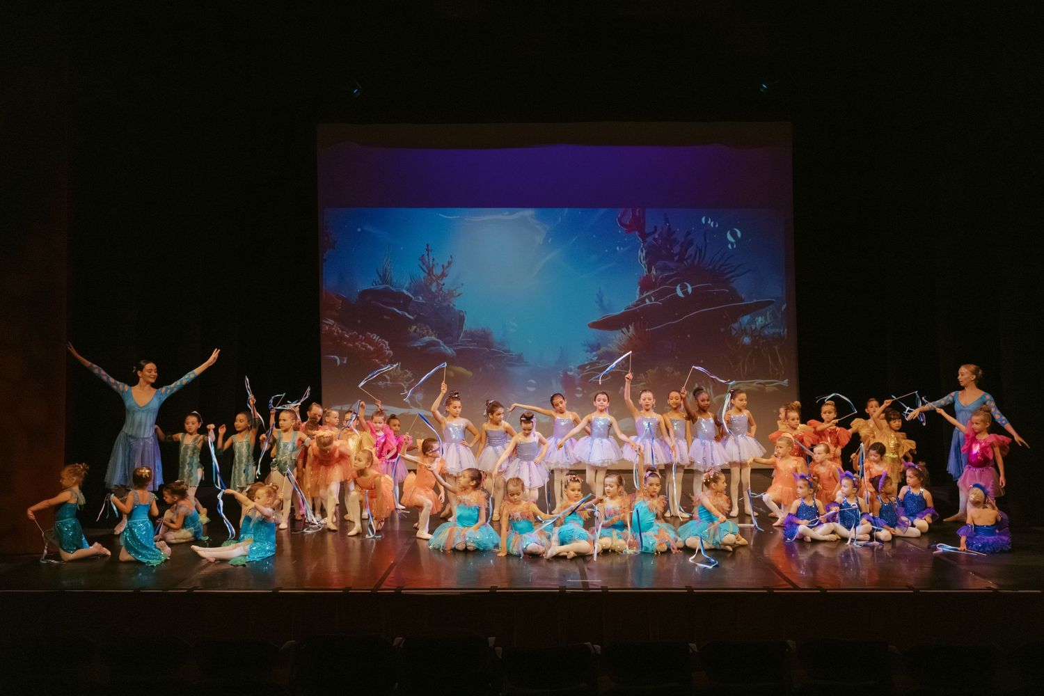 Performers on stage in colorful costumes dance during a theatrical production with blue projected backdrop scenery.