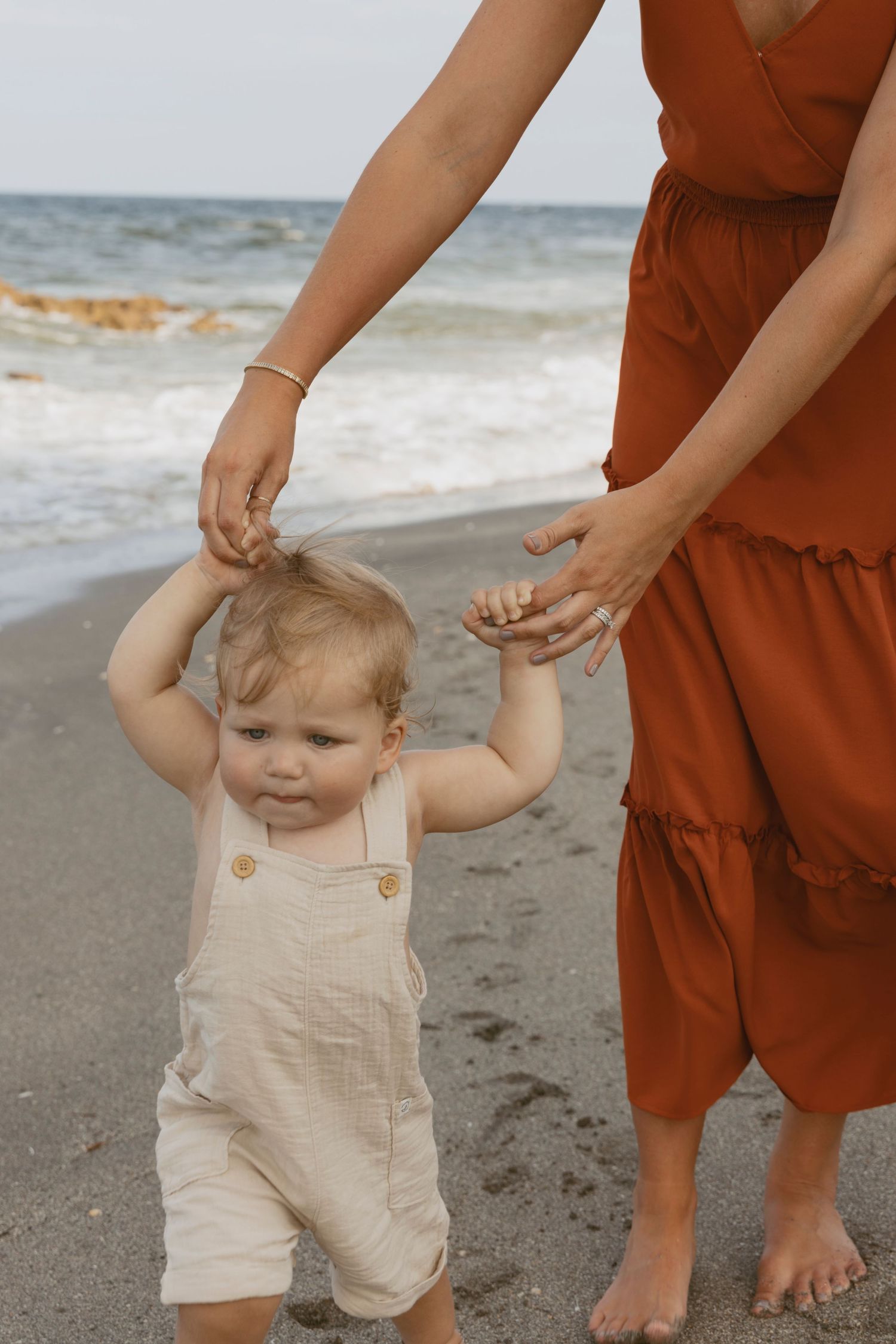 Toddler in beige overalls walking on beach while holding parent's hands.