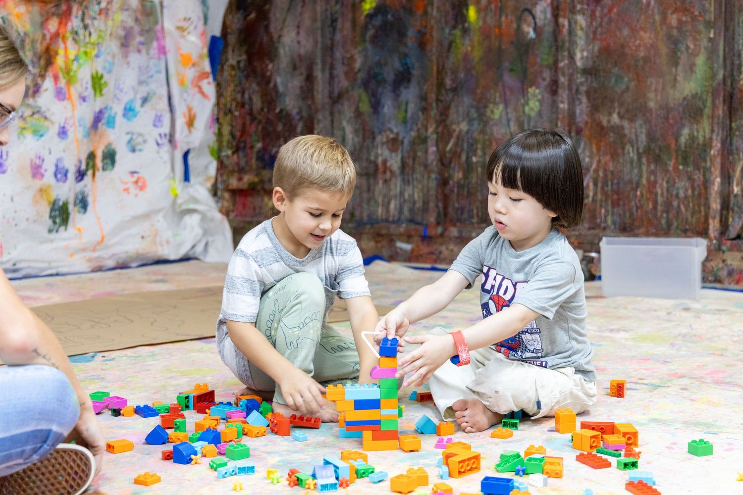 Two children play with colorful building blocks on the floor in an artistic space with paint-splattered walls.