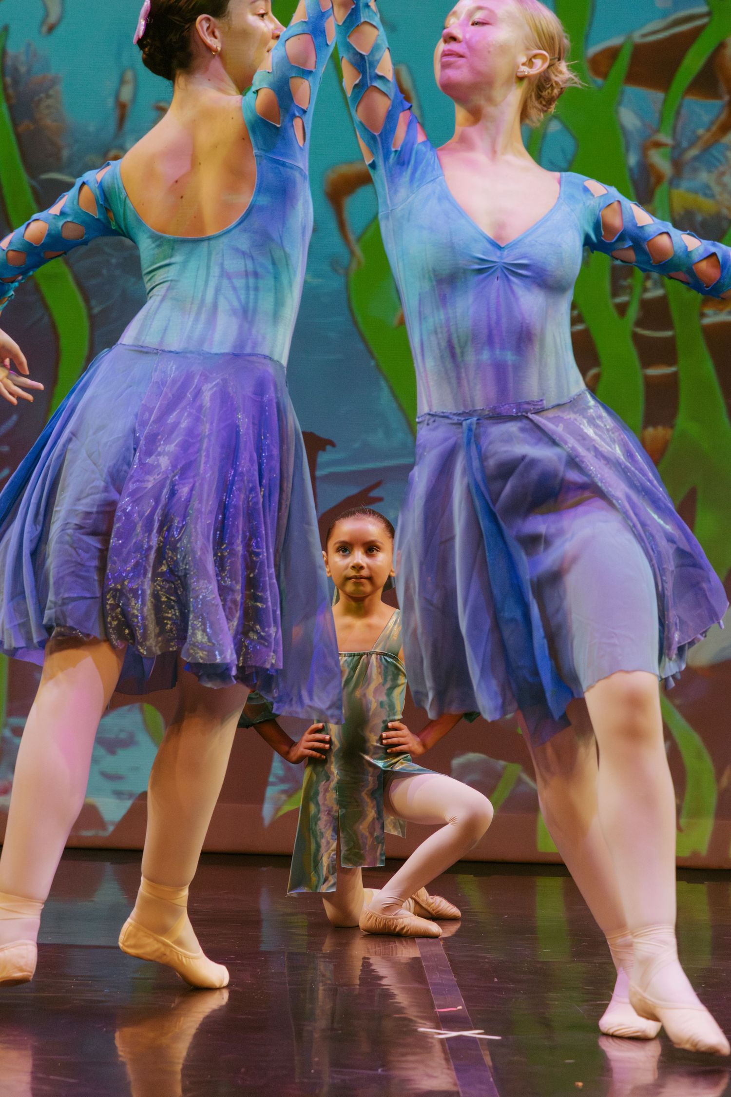 Ballet dancers perform in flowing purple and blue costumes on stage with a colorful backdrop.