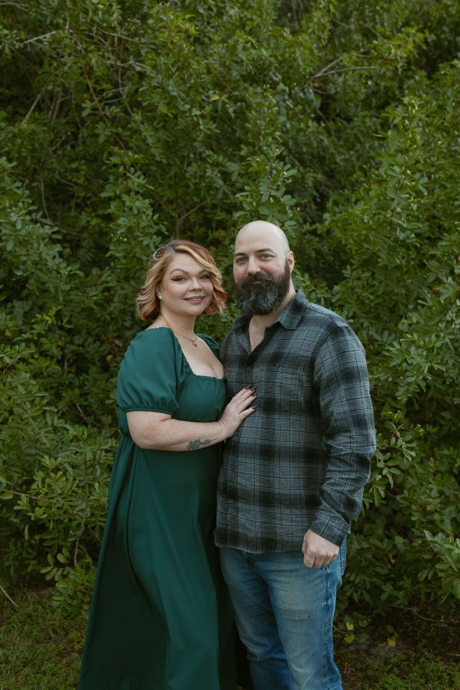 Couple poses together outdoors against lush green foliage background.