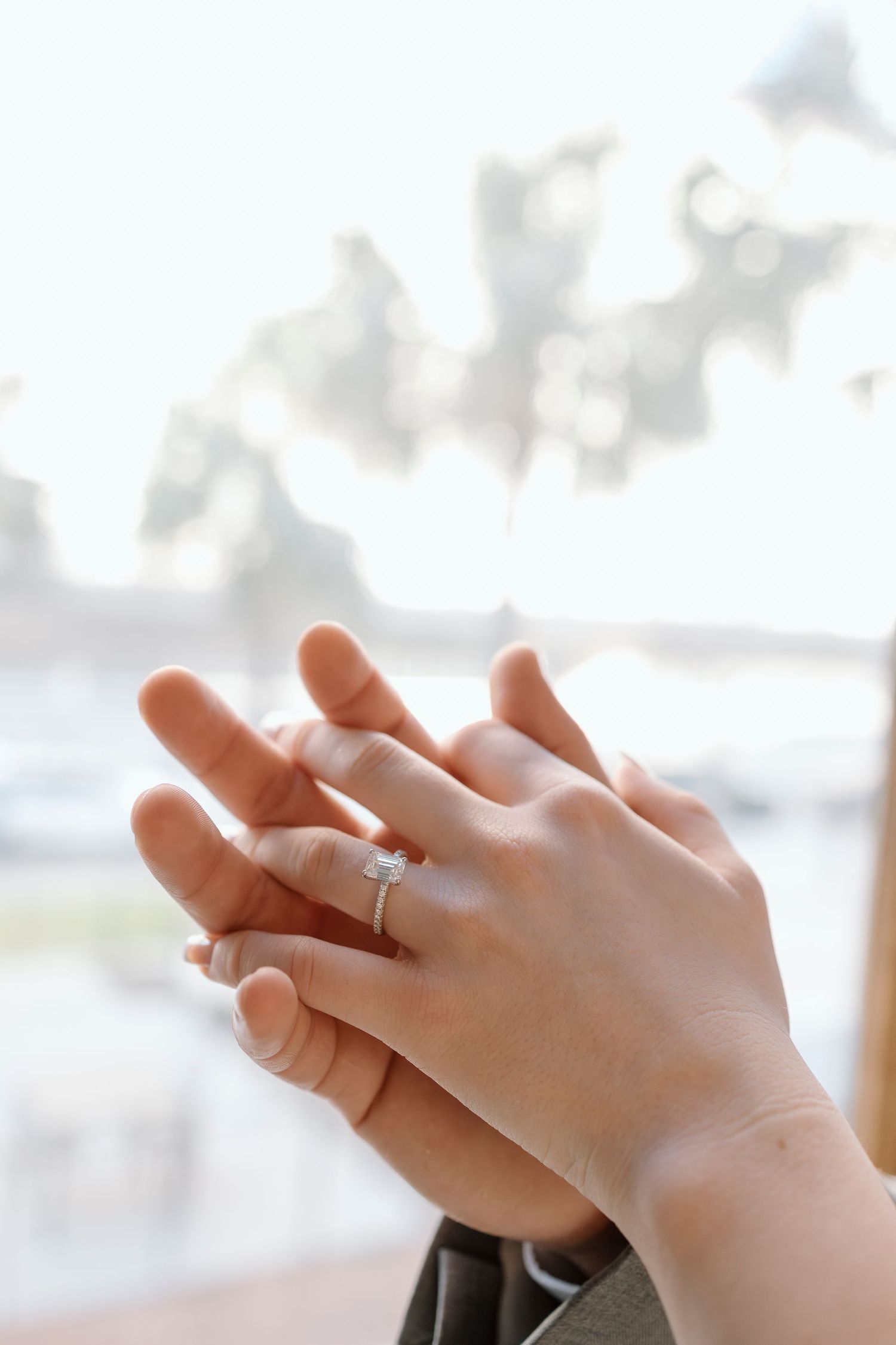 Hands with engagement ring intertwined against bright window with bokeh background.