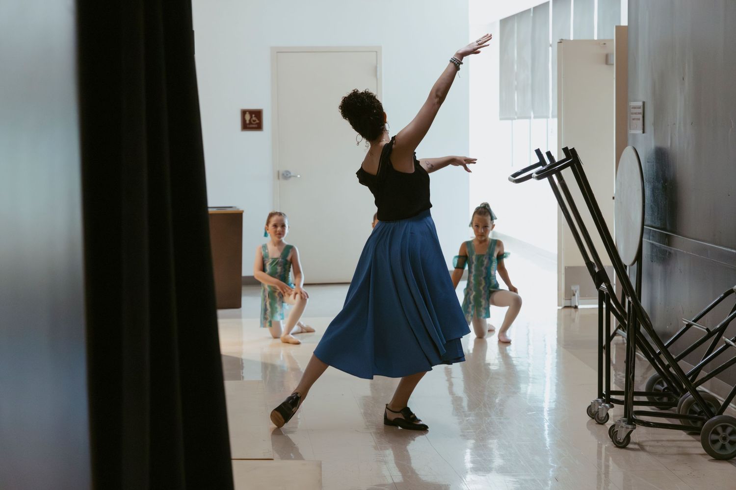 Dancers practice in a bright hallway with a silhouetted instructor in a flowing blue skirt leading the class.