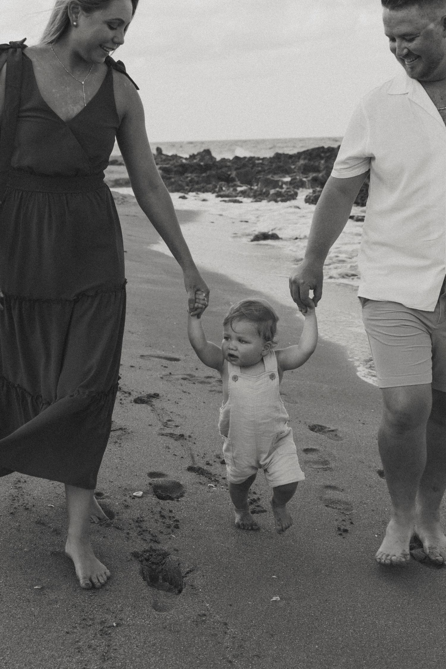Black and white beach photo of toddler walking between parents on sandy shore.