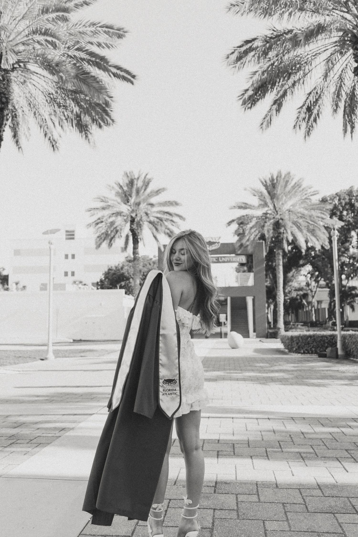 Black and white photo of graduate holding stoles while standing under palm trees.